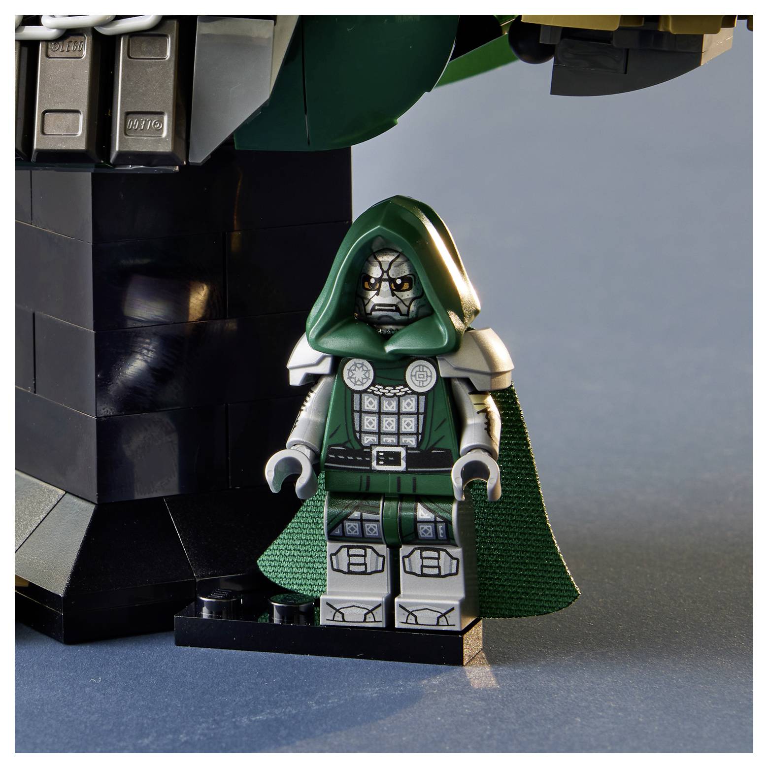 Eine LEGO Doctor Doom Minifigur in einem grünen Kapuzenumhang steht neben einem größeren schwarzen LEGO-Bauwerk auf einer blauen Oberfläche.