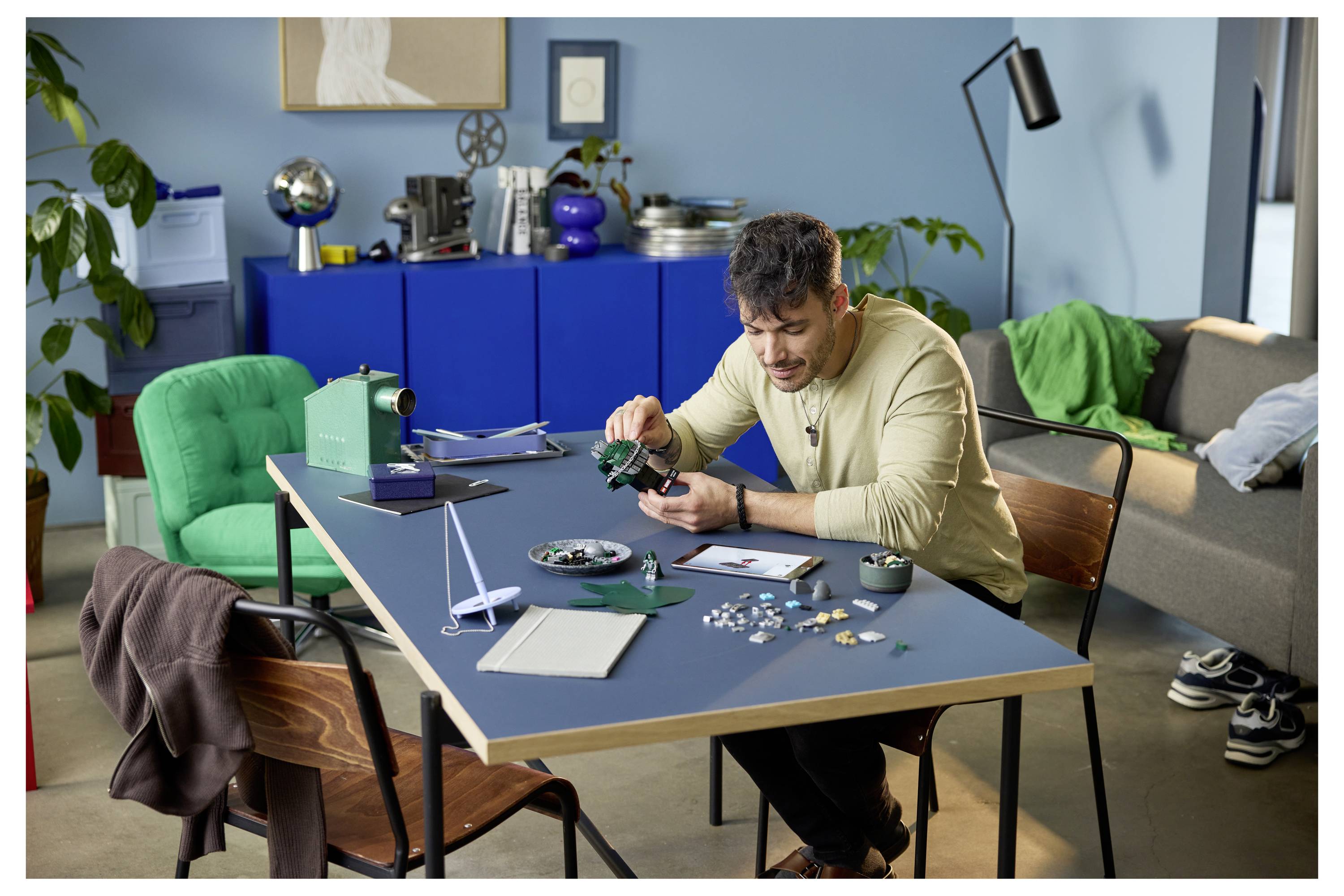 Ein Mann lehnt sich über einen Tisch und montiert sorgfältig kleine elektronische Bauteile in einem häuslichen Arbeitsbereich mit Werkzeugen, Behältern und einem Sofa im Hintergrund.