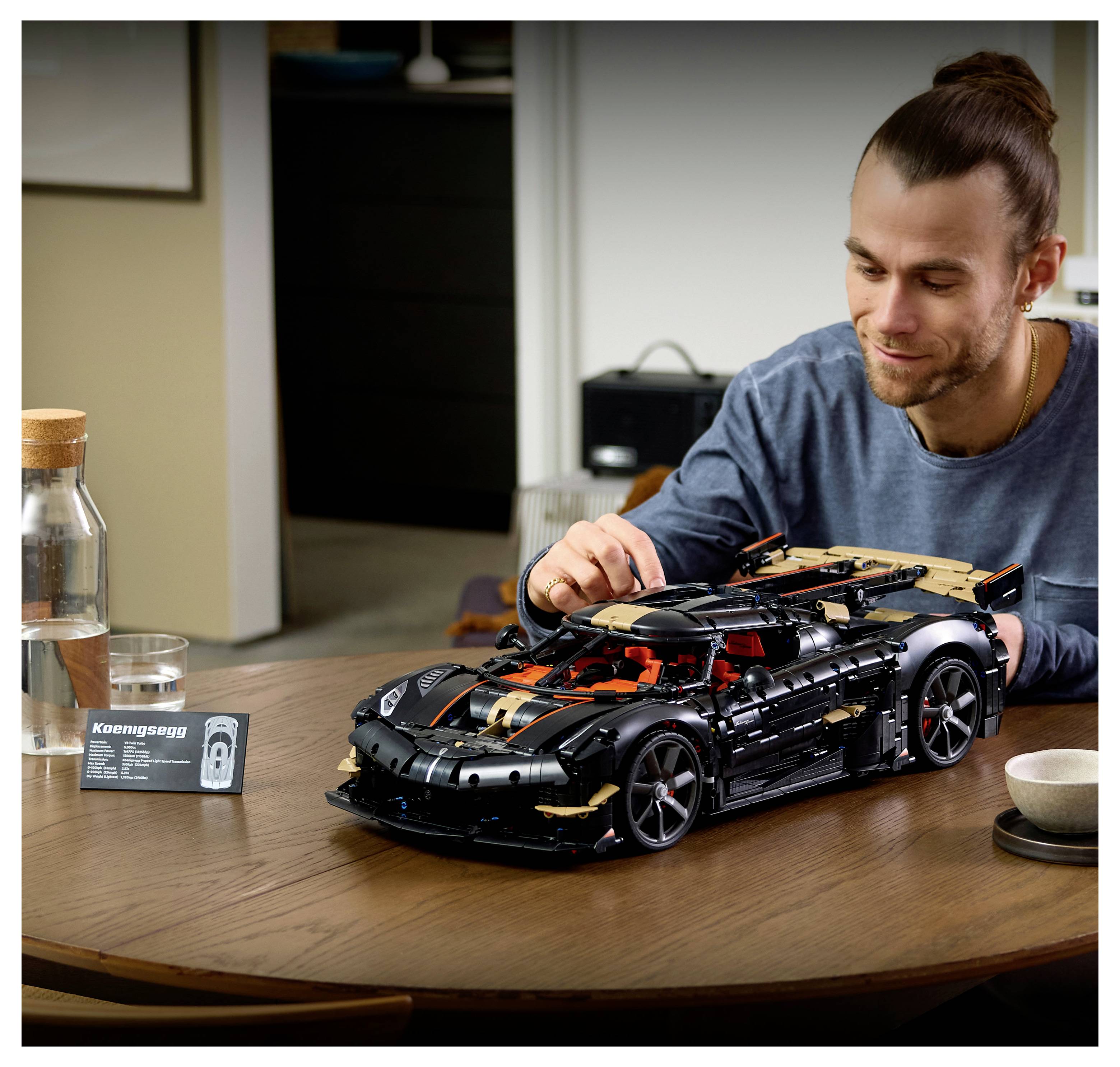 Ein Mann montiert ein detailliertes schwarzes LEGO Koenigsegg-Automodell an einem Tisch im Innenraum.
