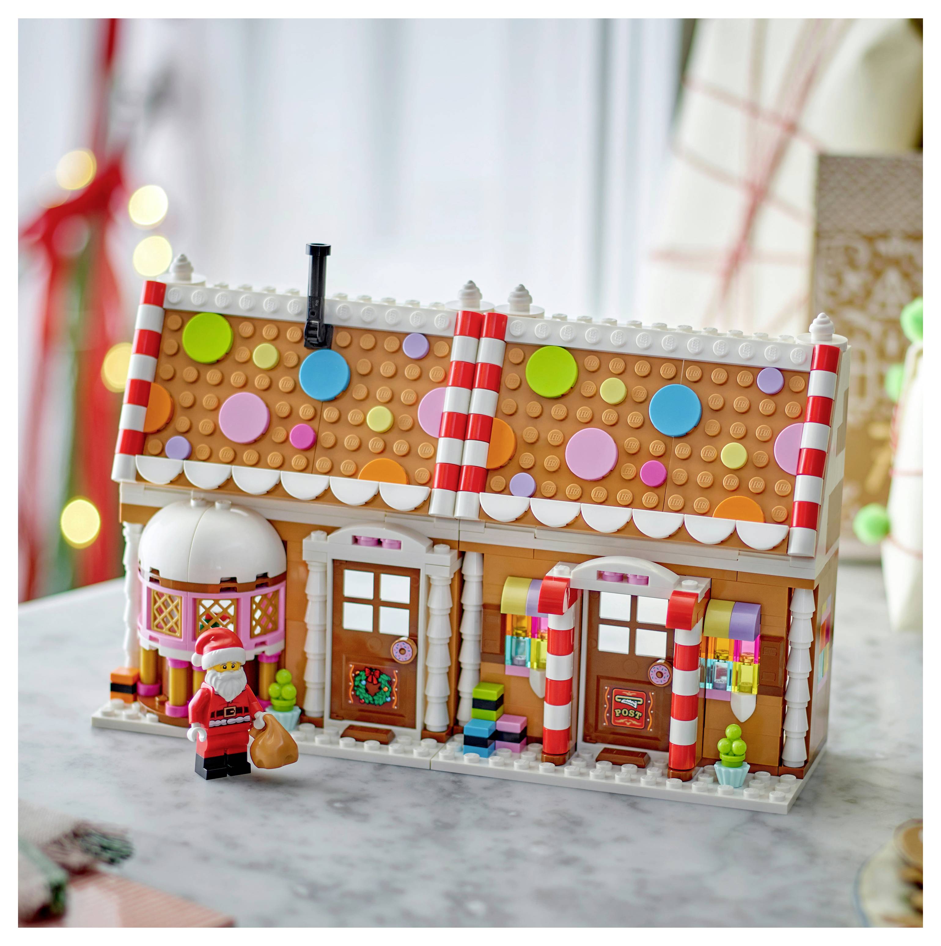 LEGO-ähnliches Lebkuchenhaus mit Süßigkeitenverzierungen und einer Santa-Minifigur, die davor auf einer Tischplatte steht.