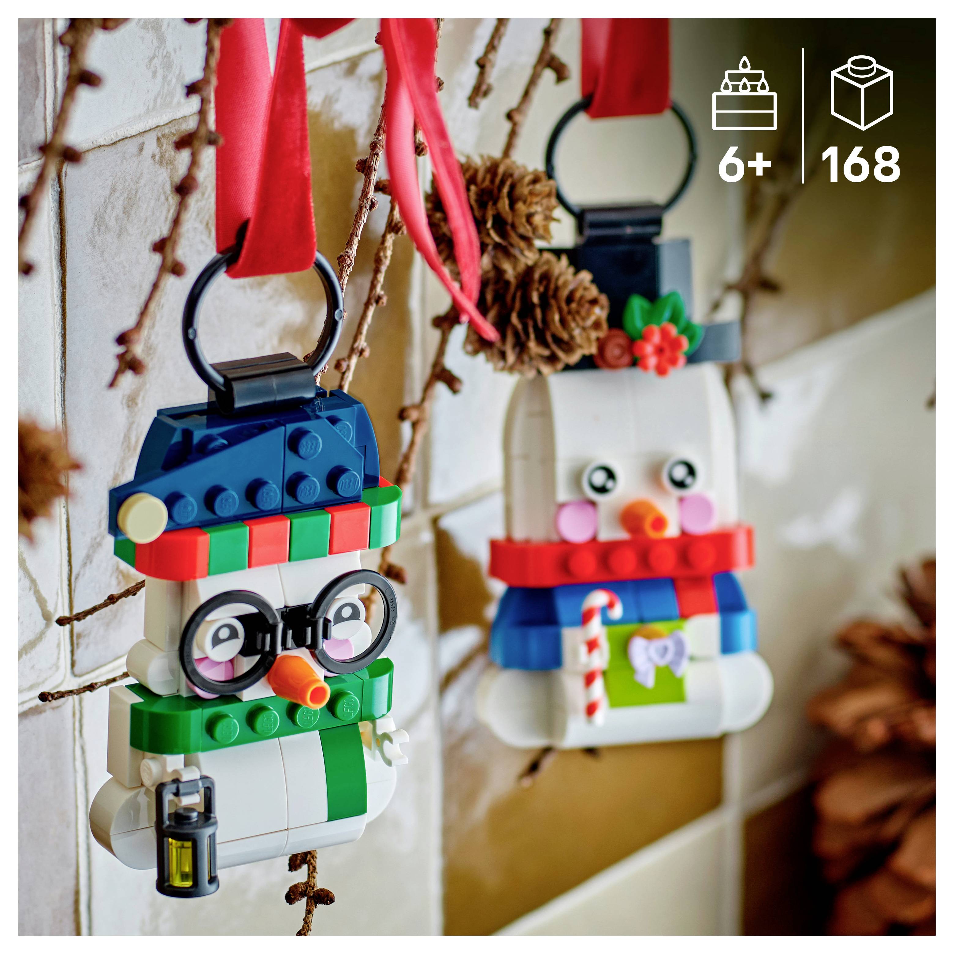 Zwei LEGO-Schneemann-Ornamente hängen an roten Bändern; einer trägt eine Brille und einen Zylinder, der andere hat einen Zuckerstab und eine Blume.