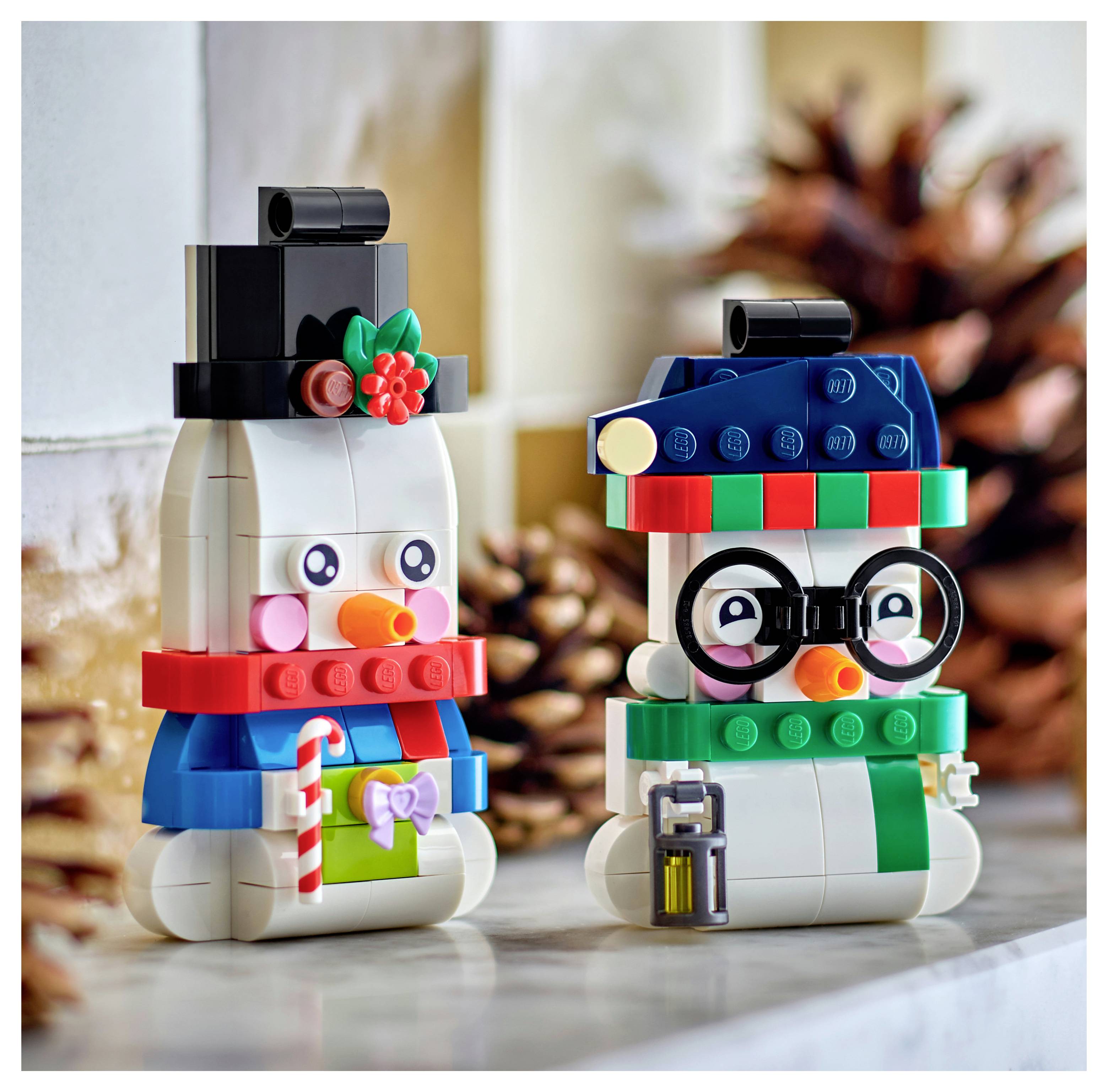 Zwei LEGO-Schneemännchen-Figuren mit Hüten, Schals und Brillen sitzen auf einer Tischplatte, wobei eine einen Zuckerstab und ein Geschenk hält, vor einem Tannenzapfen-Hintergrund.