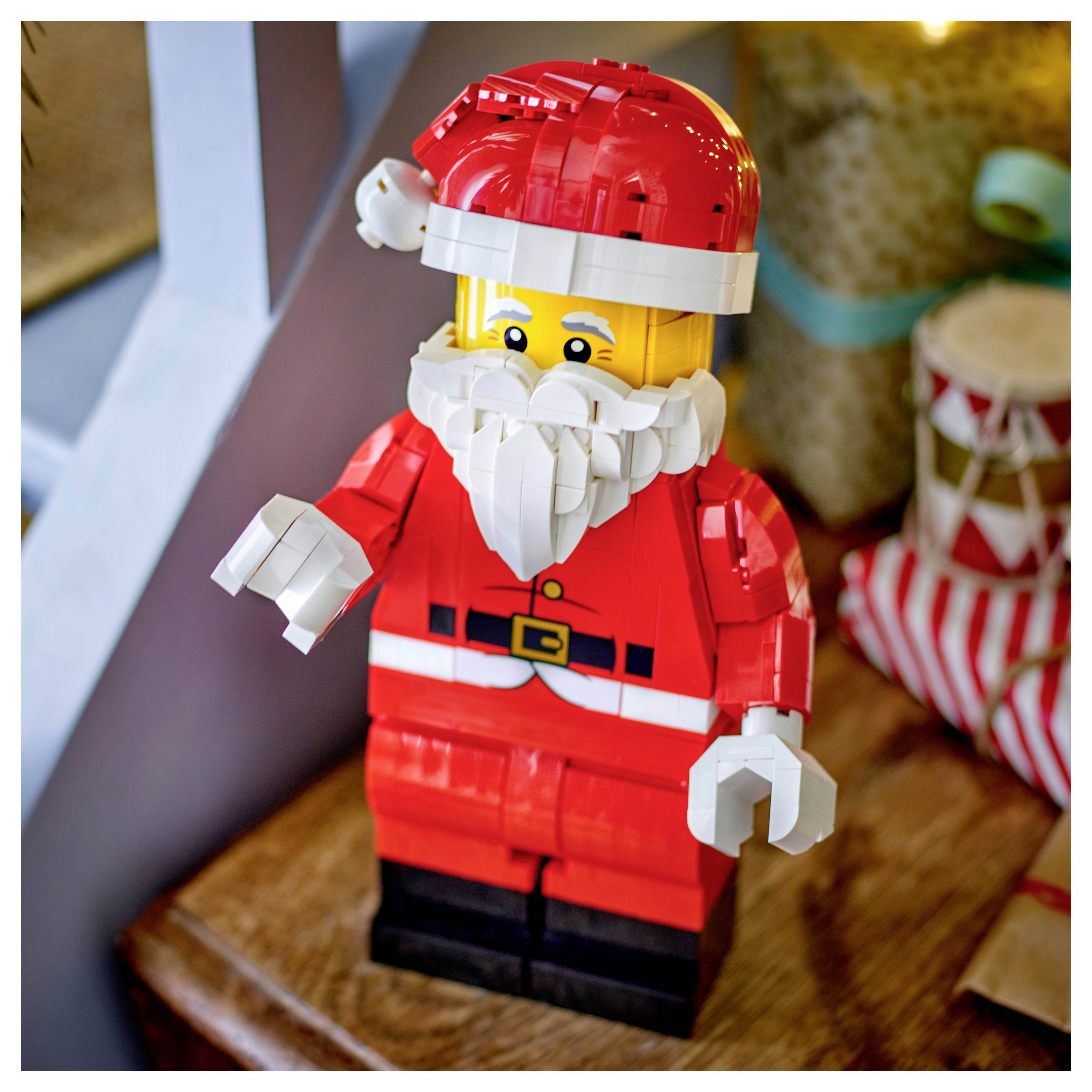 LEGO Weihnachtsmannfigur in rotem Anzug und Mütze mit weißem Bart, stehend auf einer Holzoberfläche neben einem eingewickelten Geschenk.