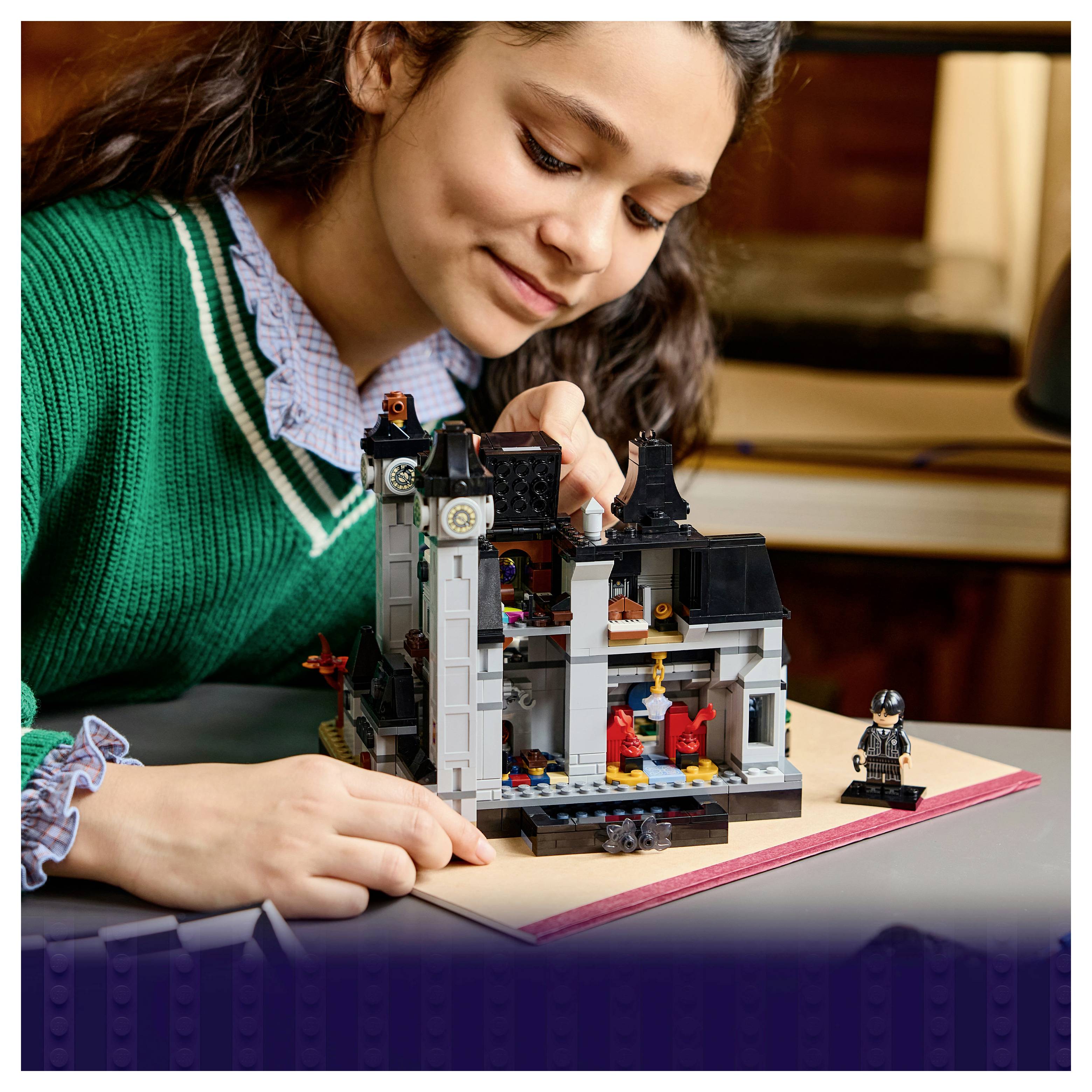 Mädchen in einem grünen Pullover baut ein detailliertes LEGO-Haus auf einem Notizbuch auf und konzentriert sich auf das Modell mit einer Minifigur daneben.