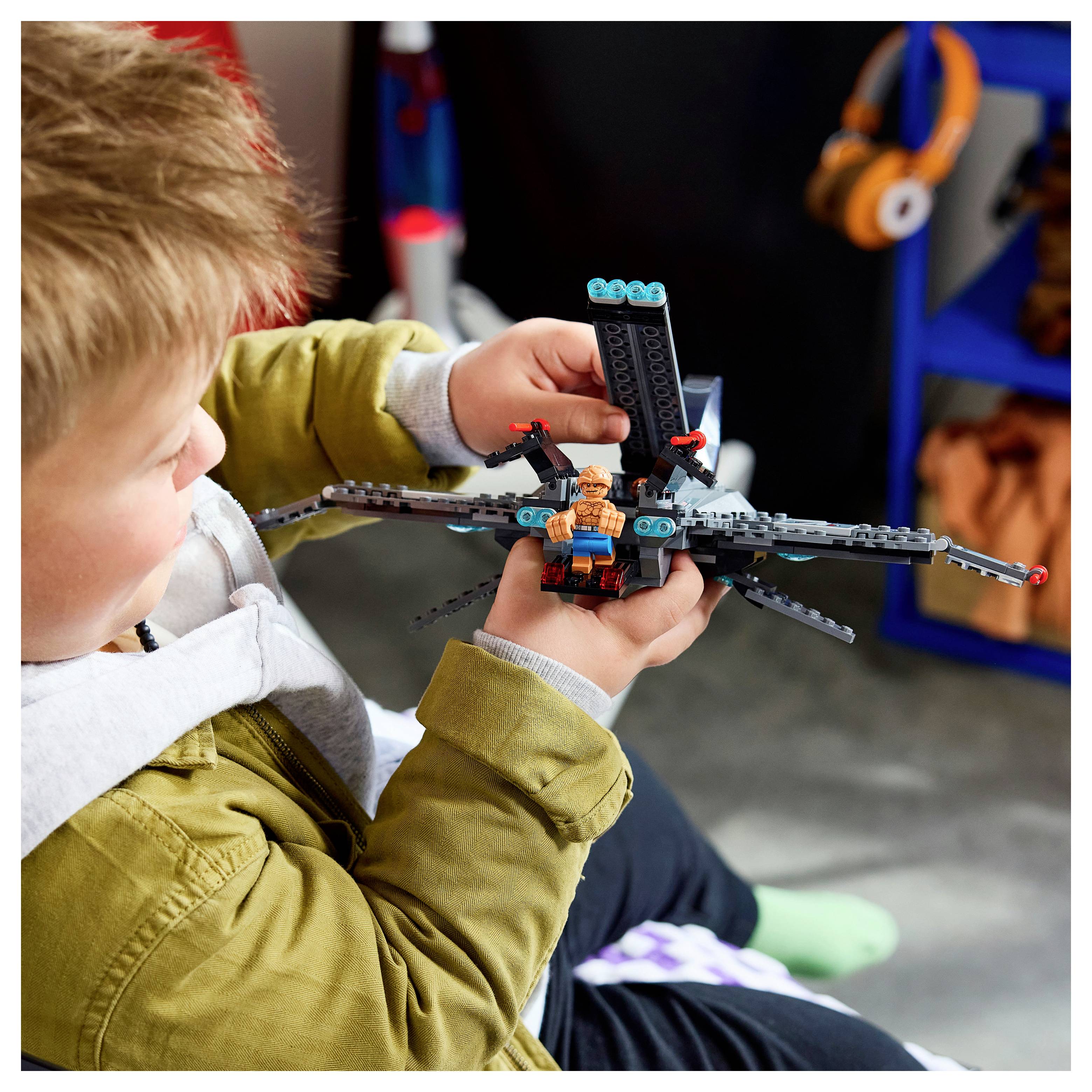 Junger Junge, der ein LEGO-ähnliches Spielzeugflugzeug oder eine Drohne mit einer Minifigur hält und inspiziert, während er drinnen sitzt und spielt.