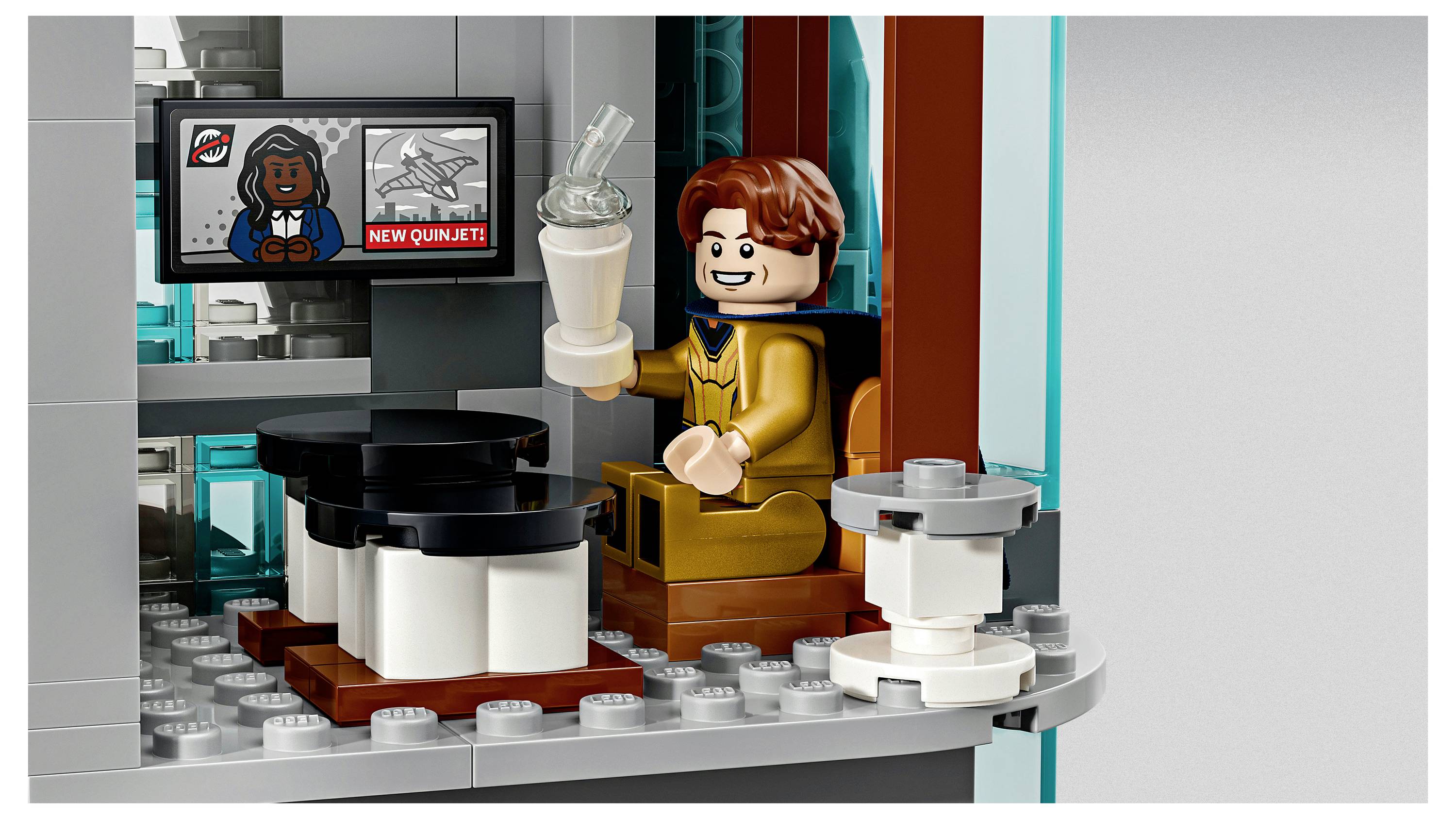 Ein LEGO-ähnliche Minifigur in einem Goldanzug sitzt an einem Schreibtisch, hält einen Milkshake in der Hand und beobachtet einen Monitor, der einen Cartoon-Avatar und den Text „NEW QUINJET" anzeigt.