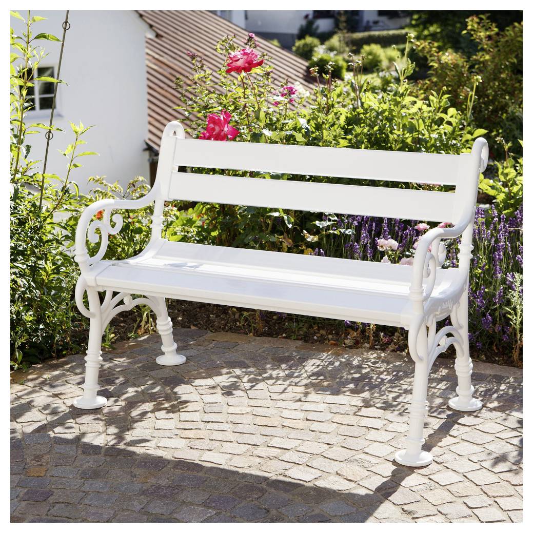 Weiße Metallgartenbank auf einer Kopfsteinpflaster-Terrasse, umgeben von blühenden Pflanzen und grünem Laub im Sonnenlicht.