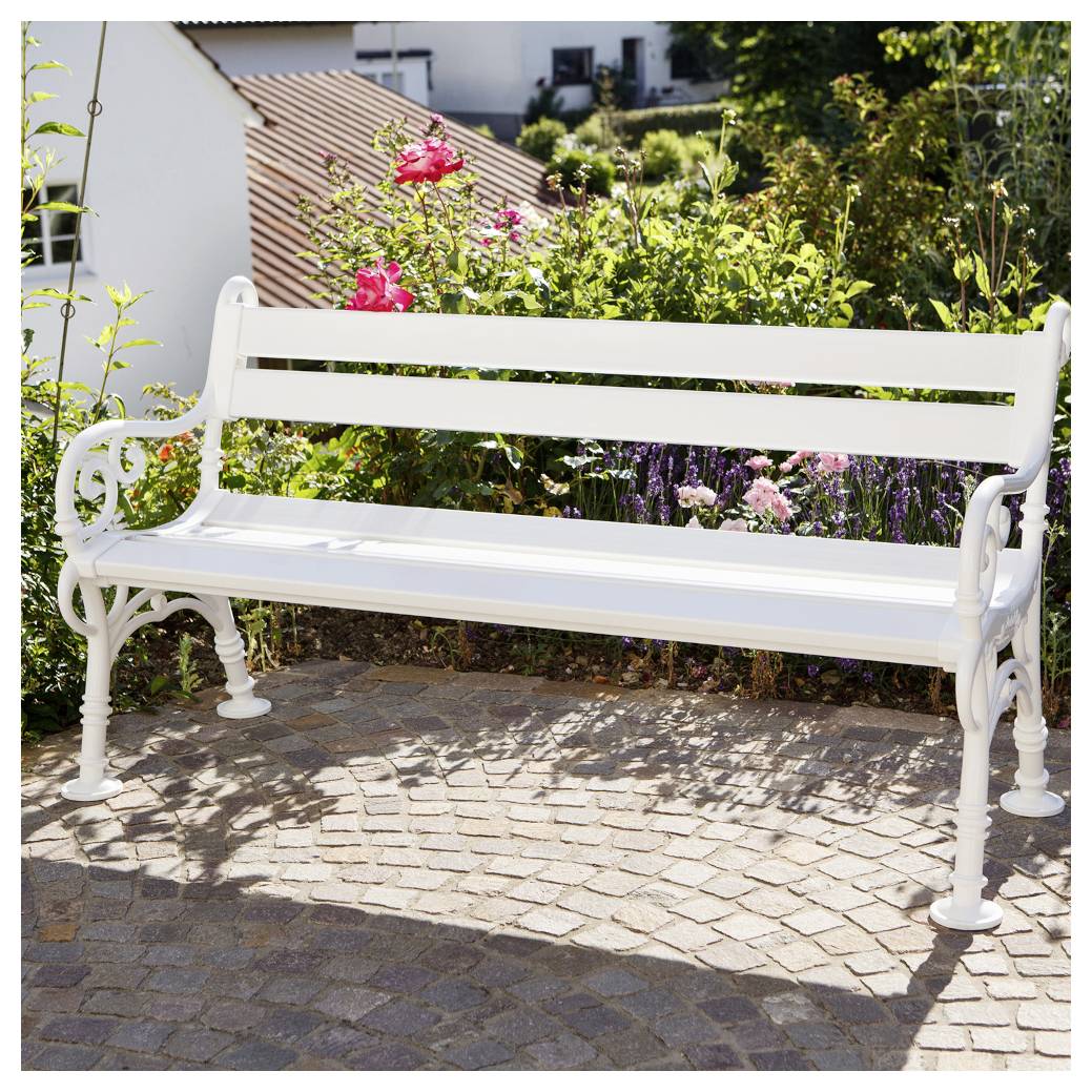 Weiße Gartenbank auf einer Kopfsteinpflaster-Terrasse vor blühenden Sträuchern und einem Haus