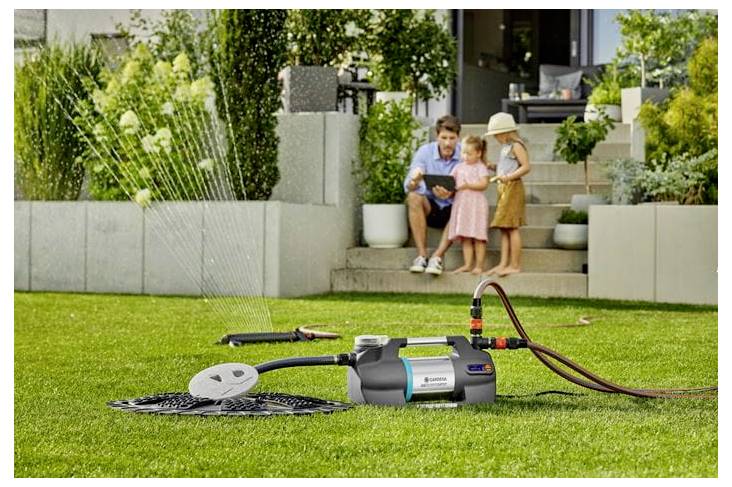 Tragbarer elektrischer Hochdruckreiniger auf einem Rasen, der Wasser sprüht, um eine Terrasse zu reinigen, während ein Mann und zwei Kinder auf den Stufen zuschauen.