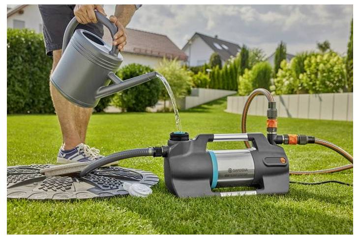 Eine Person bewässert eine Gartenschlauchpumpe auf dem Rasen und füllt eine Gießkanne, während verbundene Schläuche zu einem Haus im Hintergrund führen.