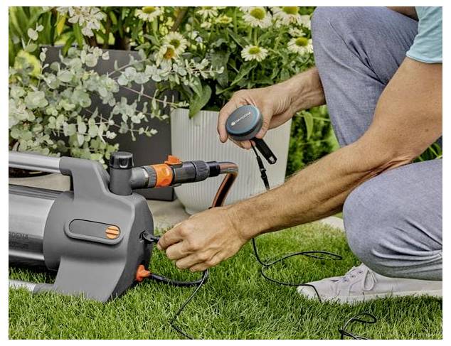 Eine Person kniet auf dem Gras und verbindet einen Schlauch und ein Stromkabel mit einem grau-orangefarbenen Gartengerät in einem Hinterhof mit Blumentöpfen.