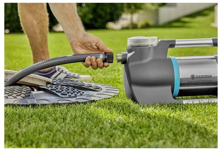 Eine Person befestigt einen Gartenschlauch an einem grauen GARDENA-Bewässerungswerkzeug auf einem Rasen, mit Gartengeräten in der Nähe.
