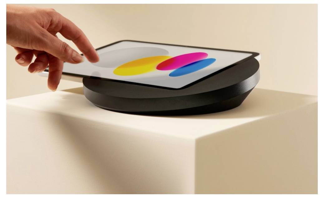 Hand greift nach einem Tablet auf einem runden Ständer, das bunte überlagernde Formen anzeigt und eine Touchinteraktion oder Design-Demo andeutet.