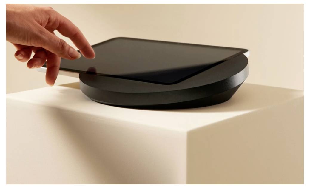 Hand schwebt über einem eleganten schwarzen kreisförmigen Gerät auf einem weißen Podest, als ob sie es gleich öffnen oder damit interagieren würde.
