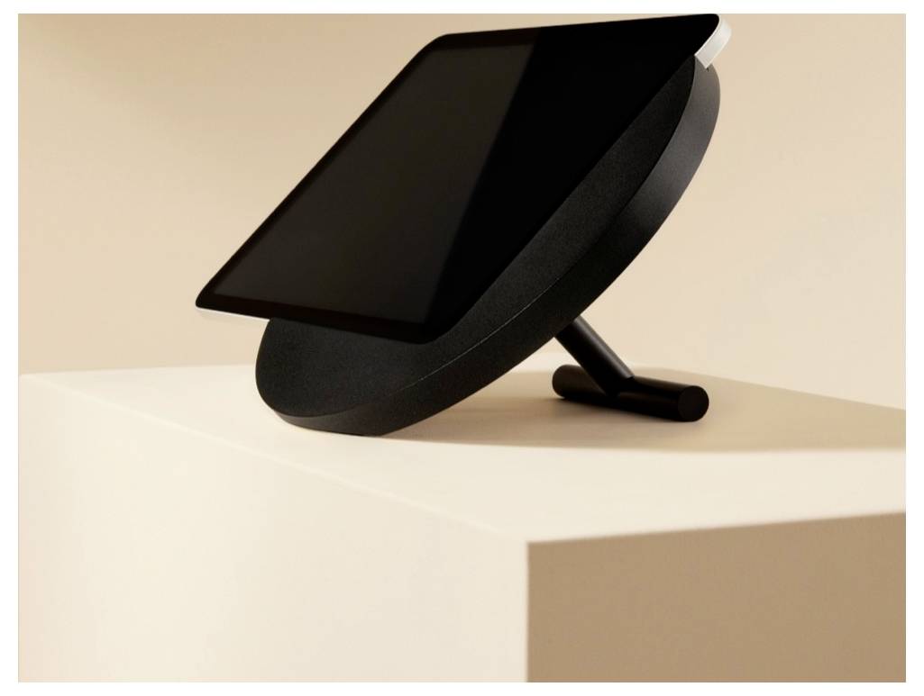 Ein schwarzes Tablet oder Display auf einem geneigten Ständer sitzt auf einem weißen Sockel vor einem beigen Hintergrund.