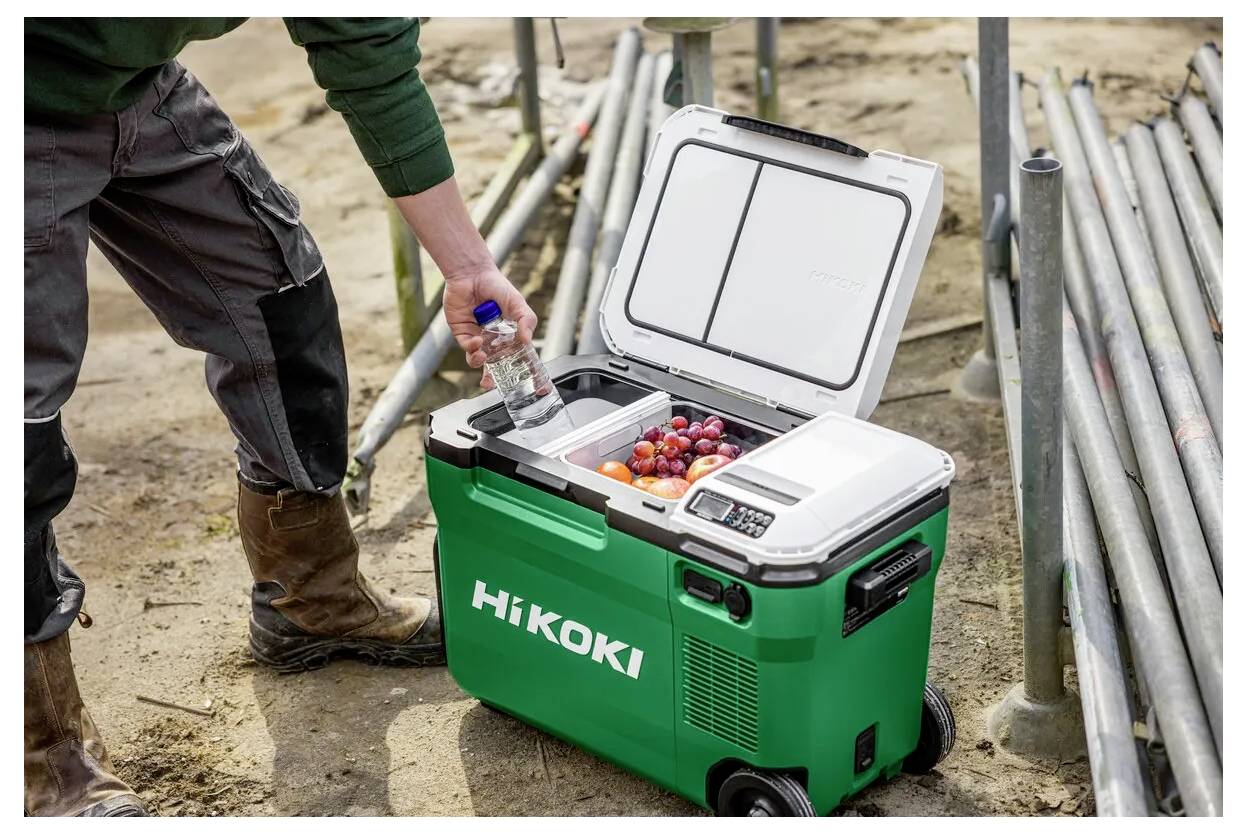 Arbeiter, der eine Wasserflasche in eine offene grüne HiKOKI-Kühlbox auf einer Baustelle stellt, mit Rohren, die in der Nähe gestapelt sind