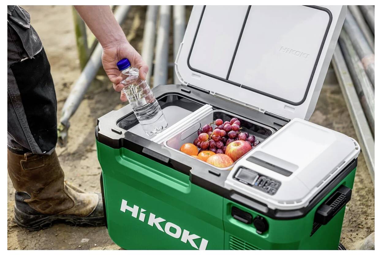 Arbeiter auf einer Baustelle stellt eine Wasserflasche in eine offene grüne HiKOKI-Kühlbox auf Rädern neben gestapelten Metallrohren ab.