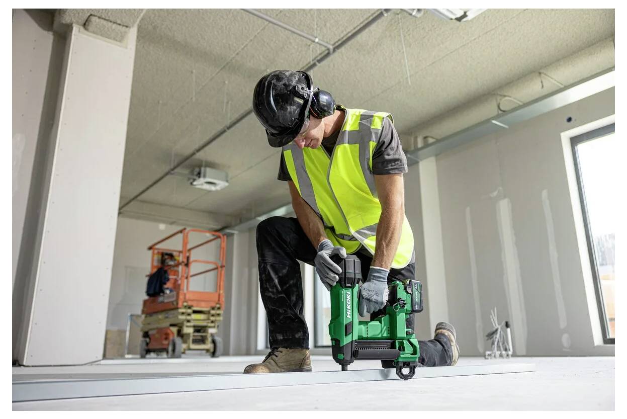 Construction worker in a safety vest and hard hat using a power tool to fasten flooring inside a building under construction.