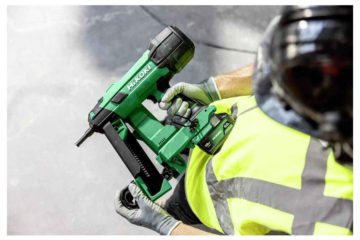 Construction worker in safety vest and gloves using a green cordless nail gun; another helmeted worker is partially visible in the foreground.