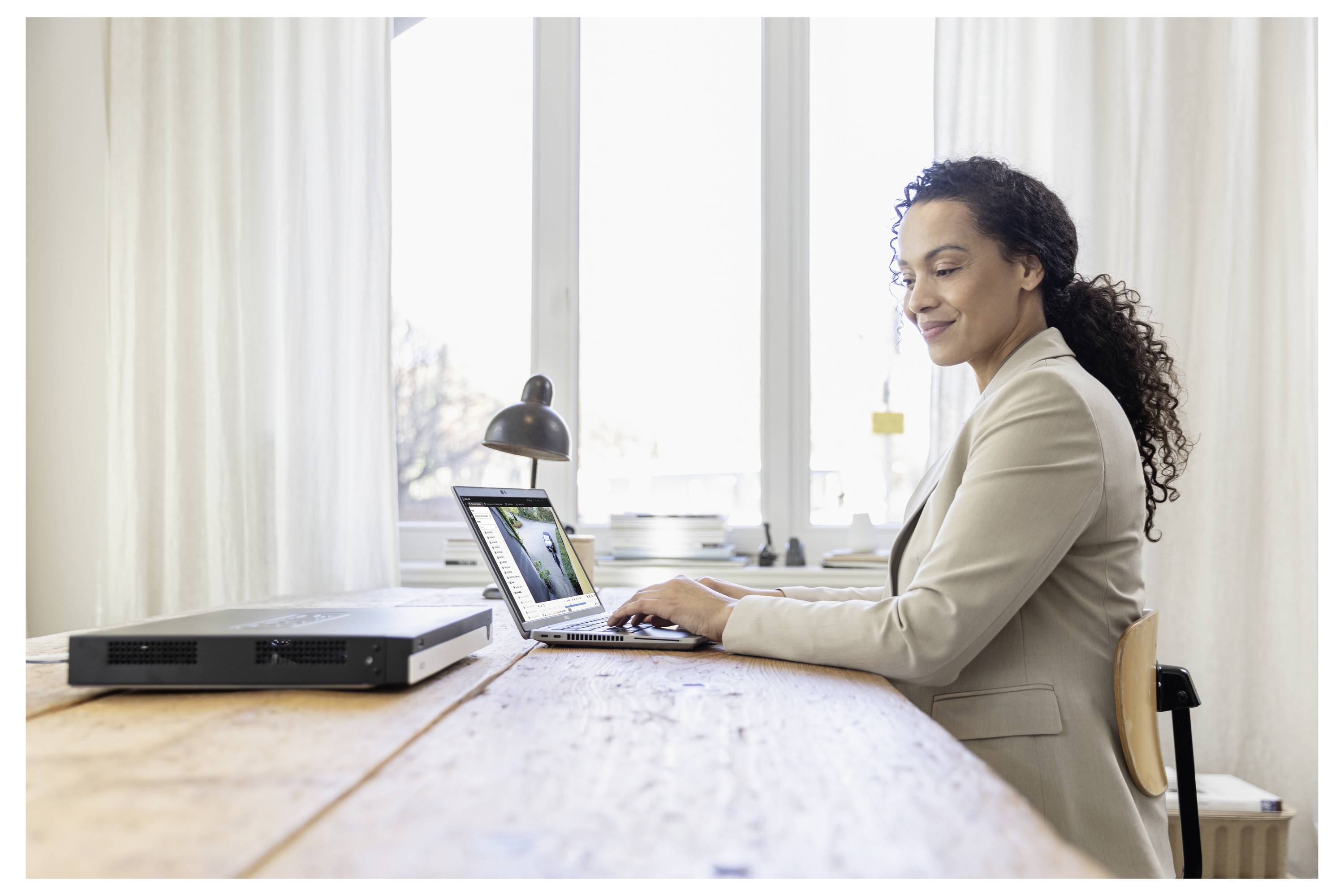 Frau in einem beigen Blazer arbeitet an einem Laptop an einem Holzschreibtisch in einem hellen Büro.