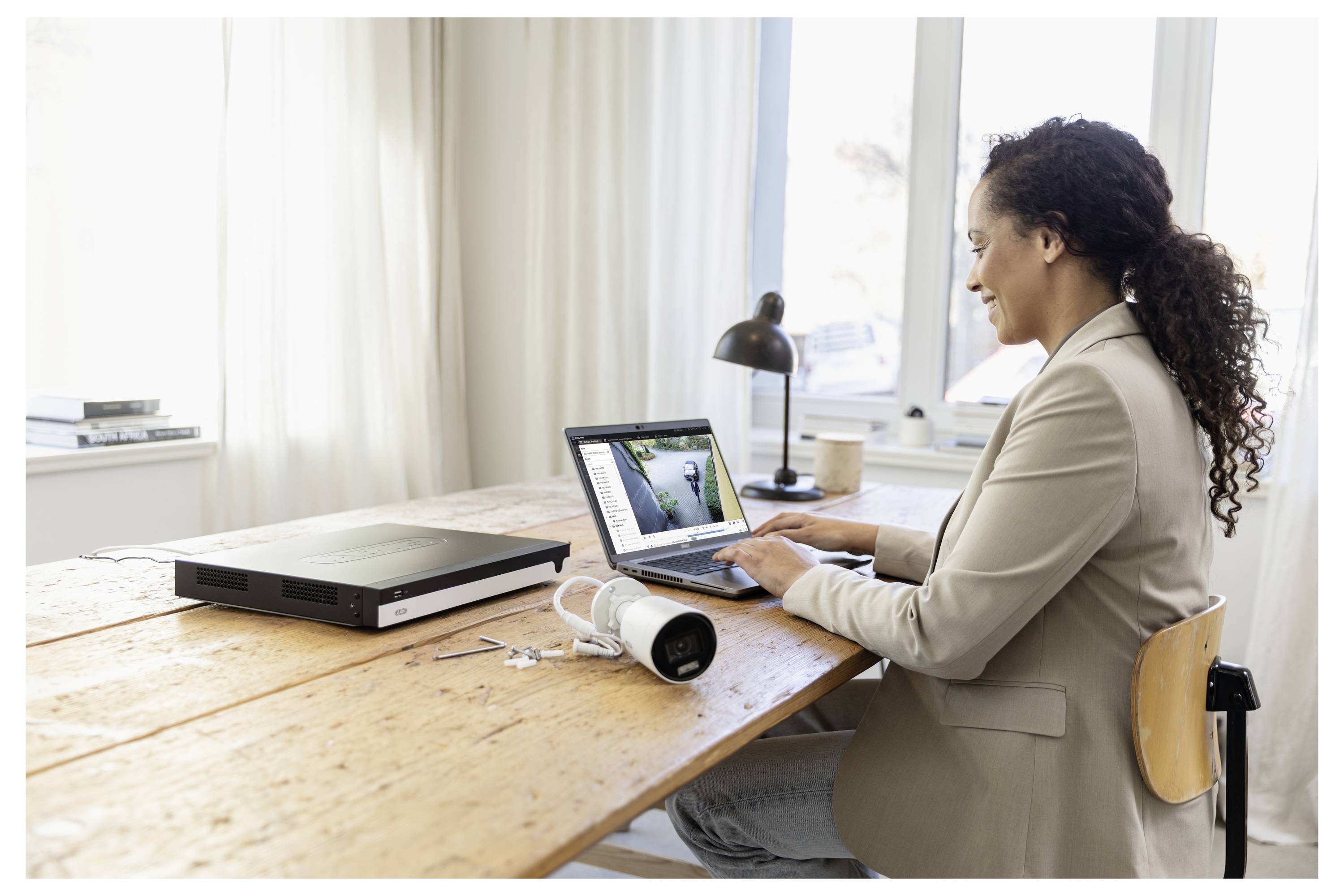 Eine Frau, die an einem Laptop an einem Holzschreibtisch in einem hellen Raum arbeitet, mit einem Drucker, einer Kaffeetasse und einer Schreibtischlampe in der Nähe.