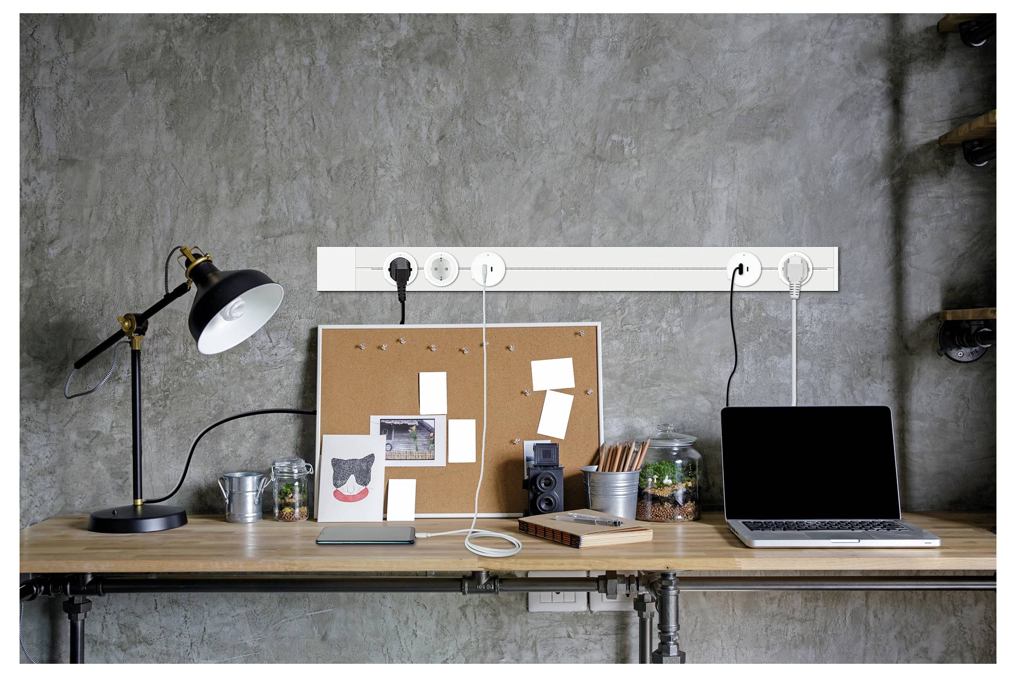 Laptop und Pinnwand auf einem Holzschreibtisch mit Lampe, Kabeln, Behältern und Kunstbedarf vor einer Betonwand, die einen kompakten Arbeitsbereich bildet.