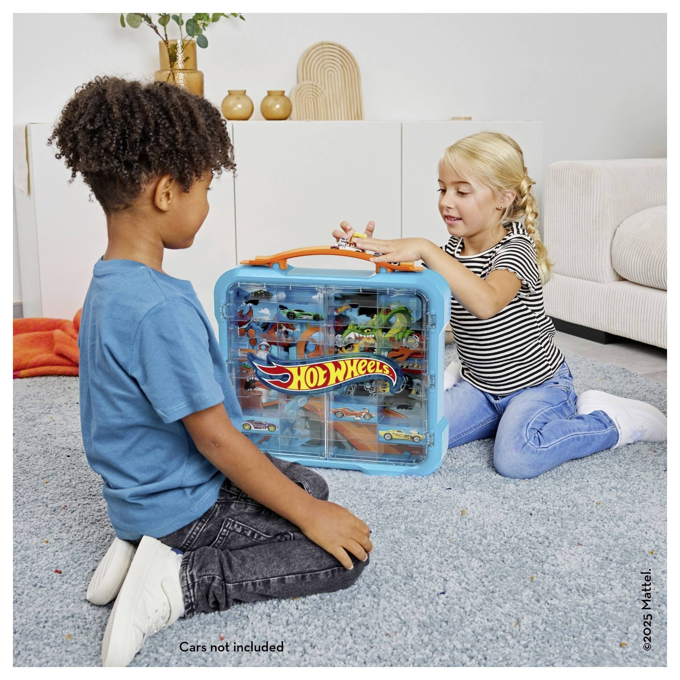 Zwei Kinder sitzen auf einem Wohnzimmerteppich und spielen mit einem blauen Hot Wheels Transportkoffer, der mit Spielzeugautos und Streckenteilen gefüllt ist.