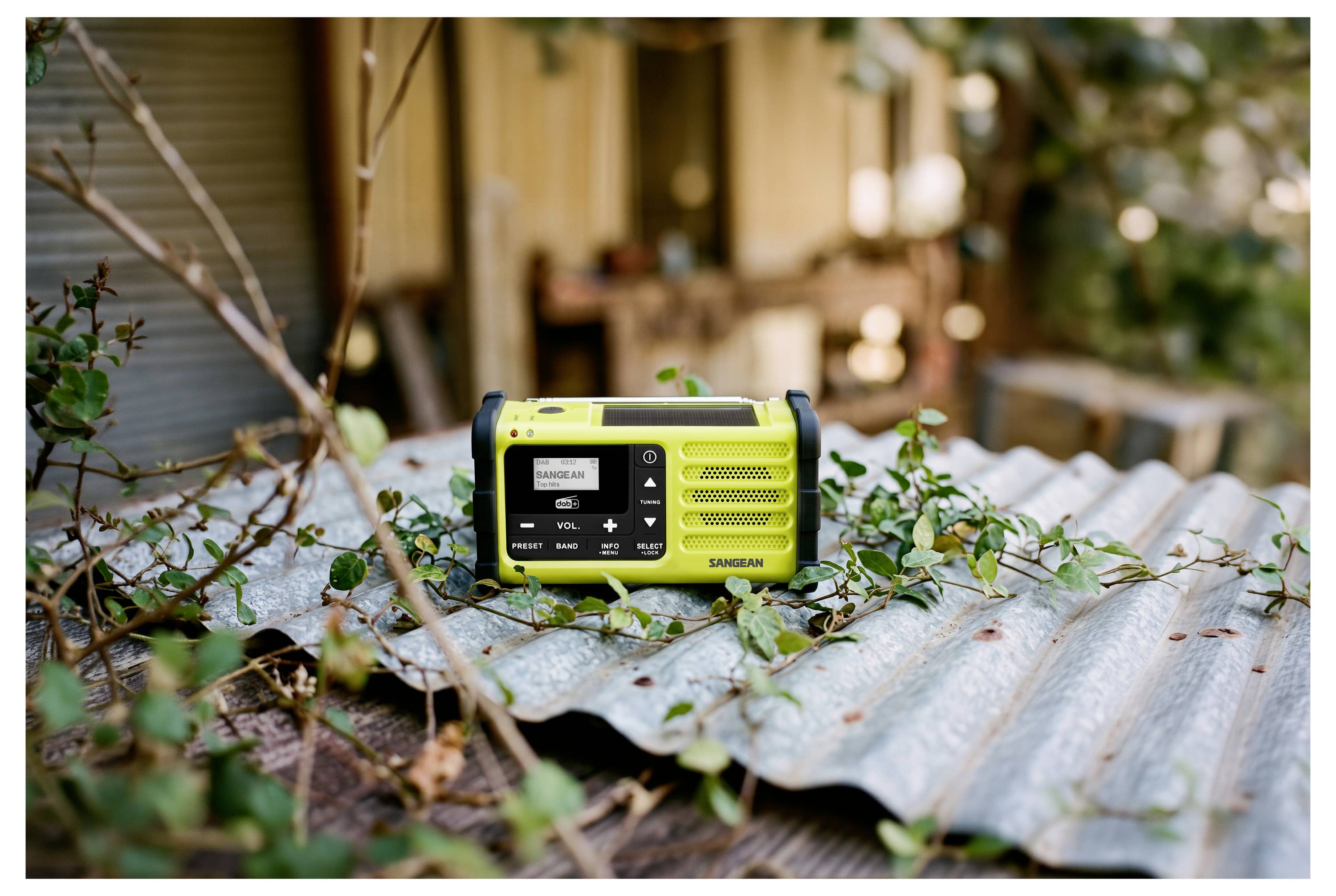 Gelbes tragbares Radio, das auf gewelltem Metall in einer Außengarten- oder Terrassenumgebung sitzt, mit Pflanzen und einem unscharfen Gebäude im Hintergrund.