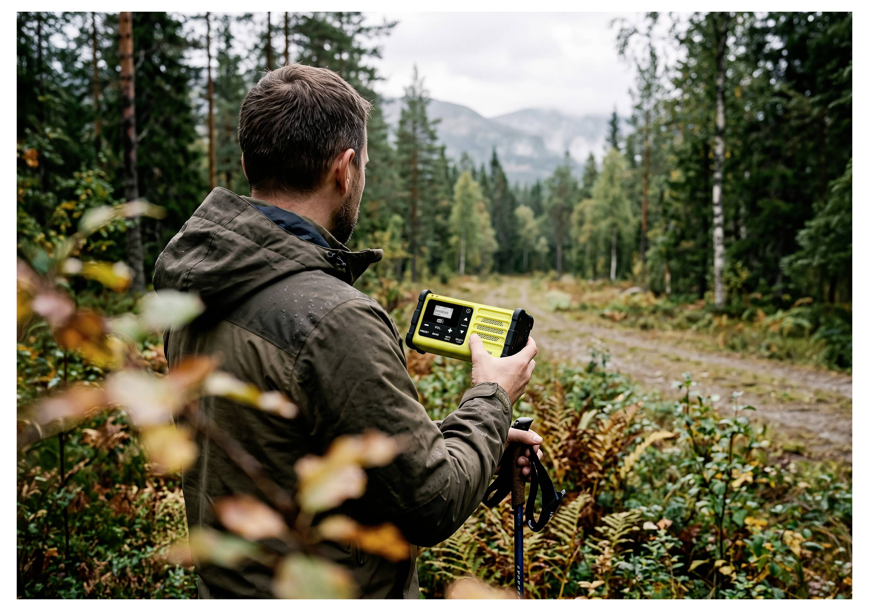 Ein Mann benutzt ein GPS-Gerät und Trekkingstöcke in einer Waldlichtung, während er einem Schotterweg zugewandt ist und entfernte Berge im Hintergrund sichtbar sind.
