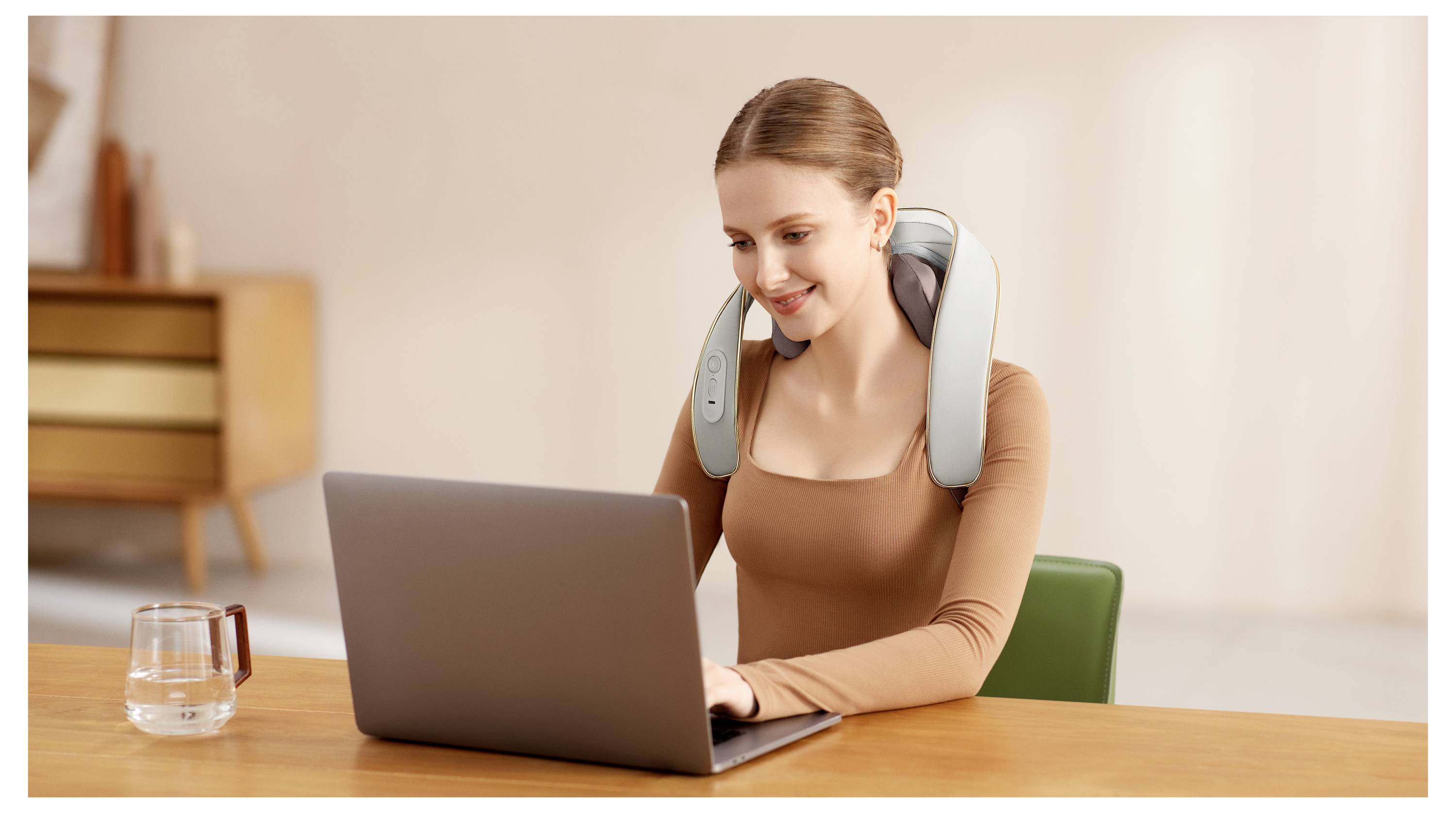 Frau, die einen Nackenmassager trägt, nutzt einen Laptop an einem Schreibtisch in einer häuslichen Umgebung, mit einem Glas Wasser in der Nähe.