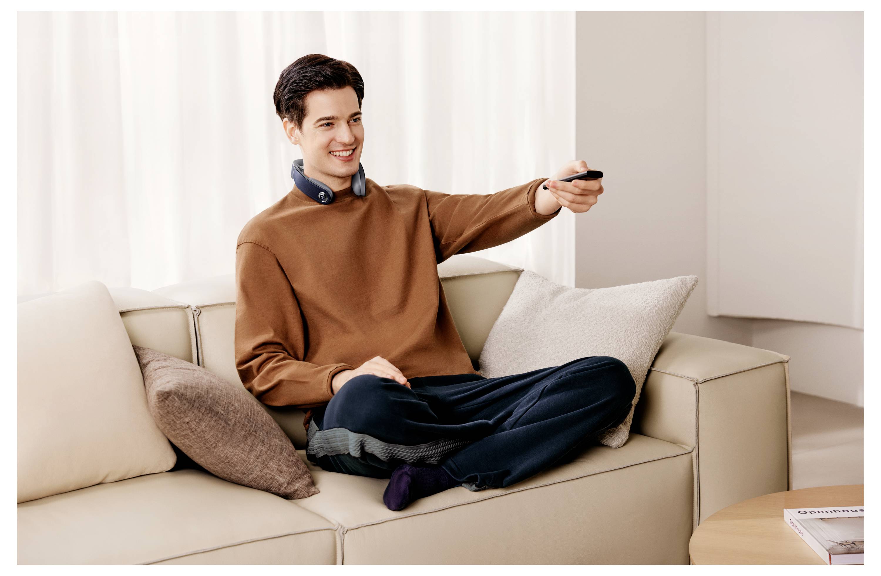 Junger Mann, der auf einem beigen Sofa sitzt, lächelt und zeigt mit einer Fernbedienung auf den Bildschirm in einem hellen Wohnzimmer.