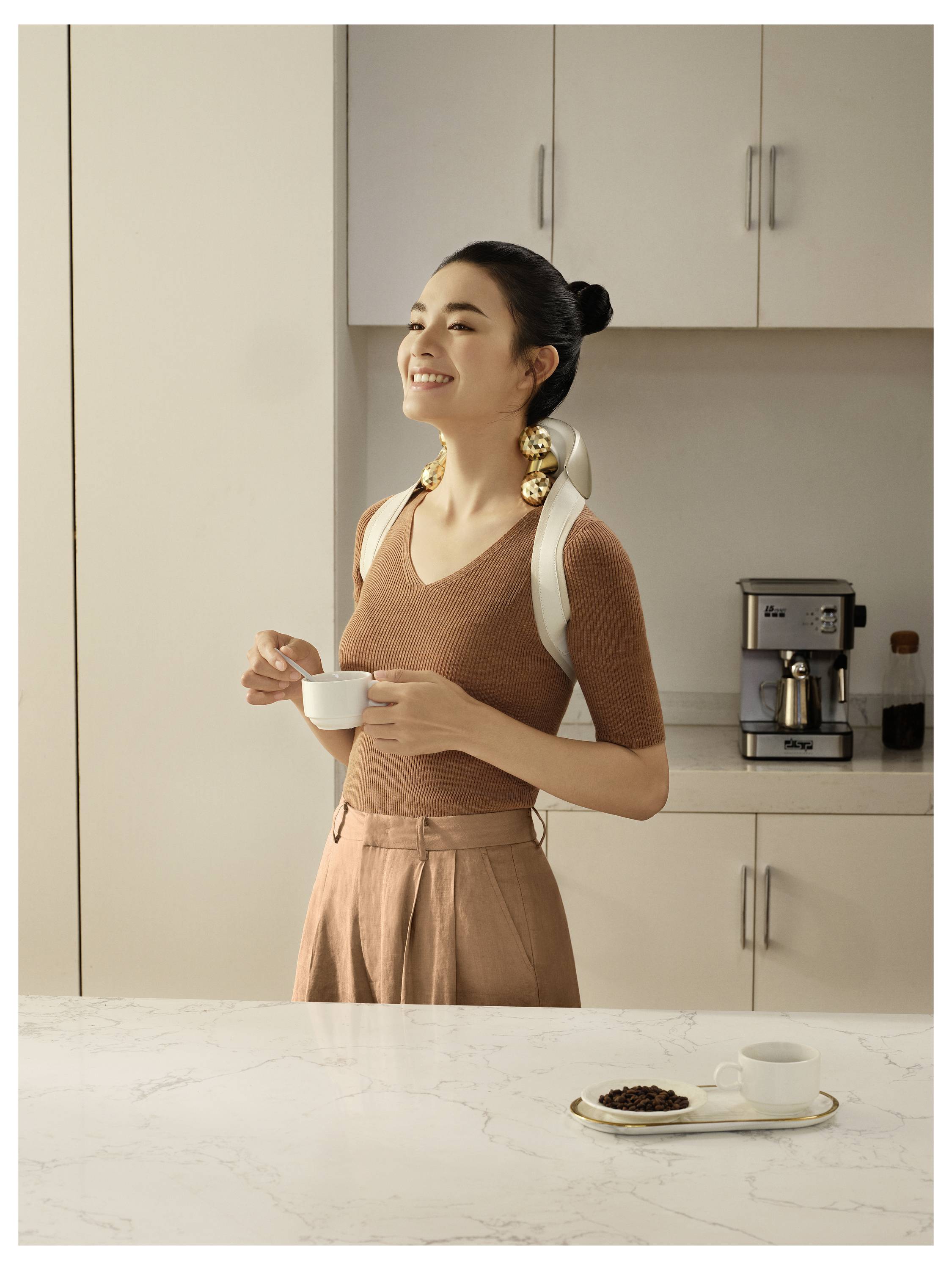 Frau in einer Küche lächelnd, während sie eine kleine Tasse hält, mit einer Kaffeemaschine und einem Teller auf der Theke hinter ihr.