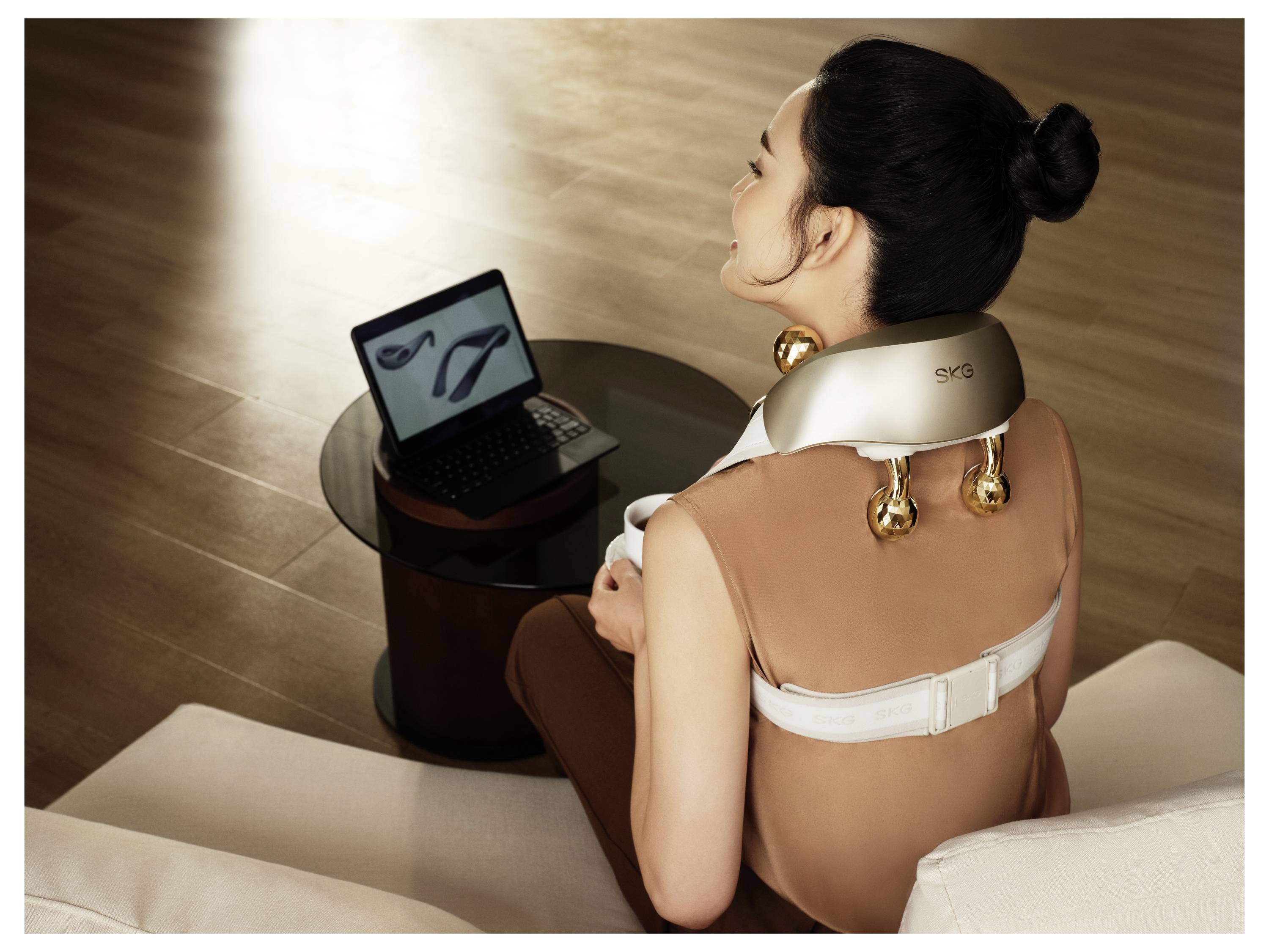 Eine Frau, die ein Nacken- und Schultermassagegerät trägt, sitzt auf einem Sofa und schaut auf einen Laptop auf einem Beistelltisch.