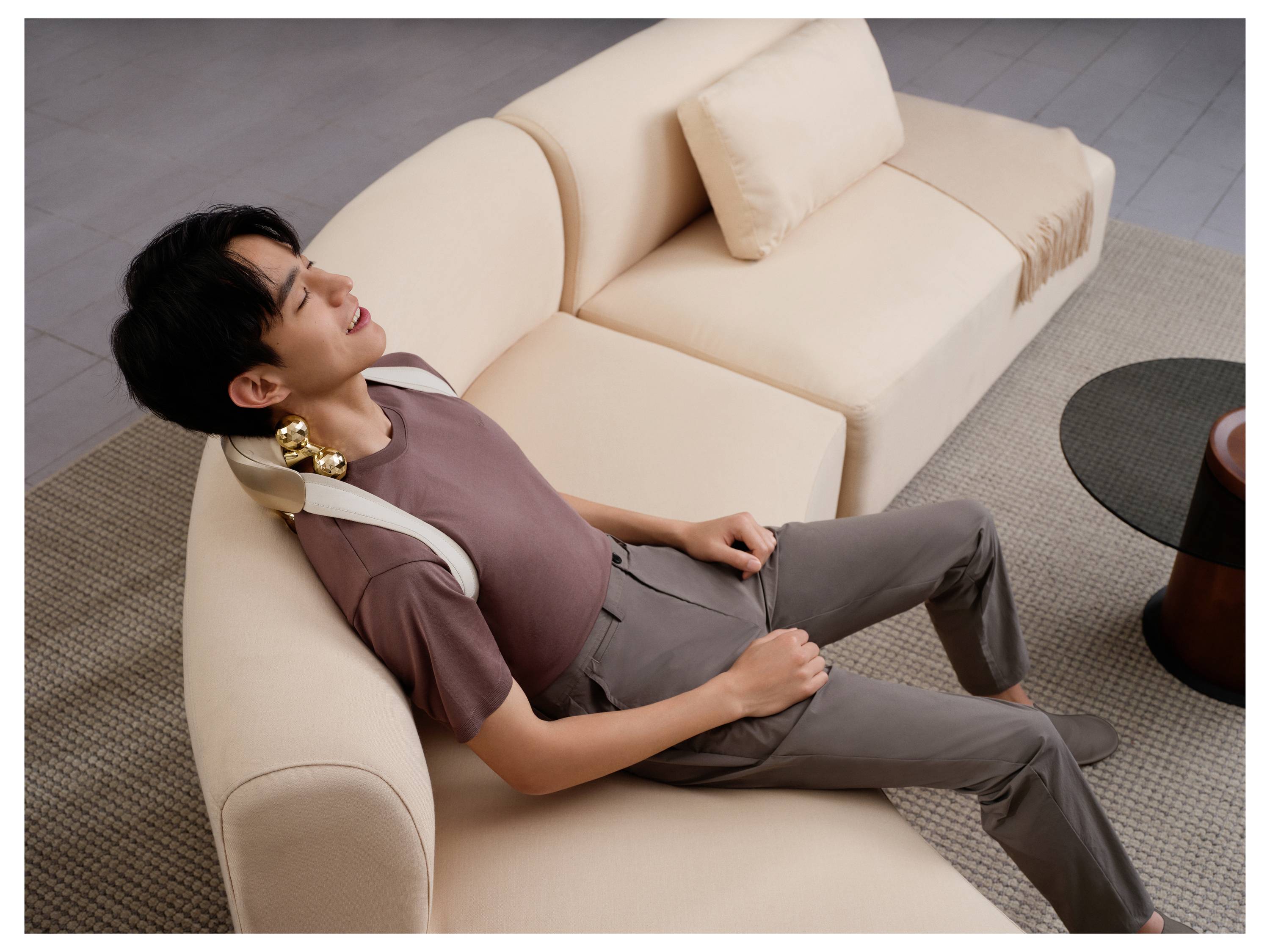 Eine Person liegt auf einem beigen Sofa und trägt ein Nackenmassagegerät, ruht sich aus mit geschlossenen Augen in einem Wohnzimmer.