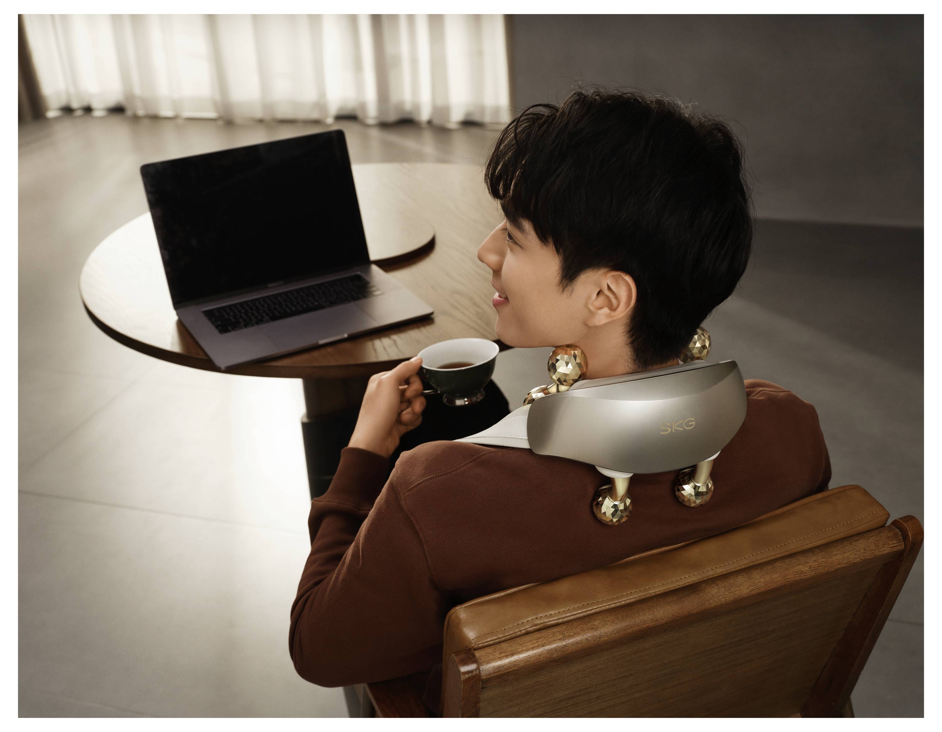 Person sitzt in einem Stuhl mit einem Nackenmassagegerät über den Schultern, hält eine Tasse und blickt auf einen Laptop auf einem kleinen Tisch.
