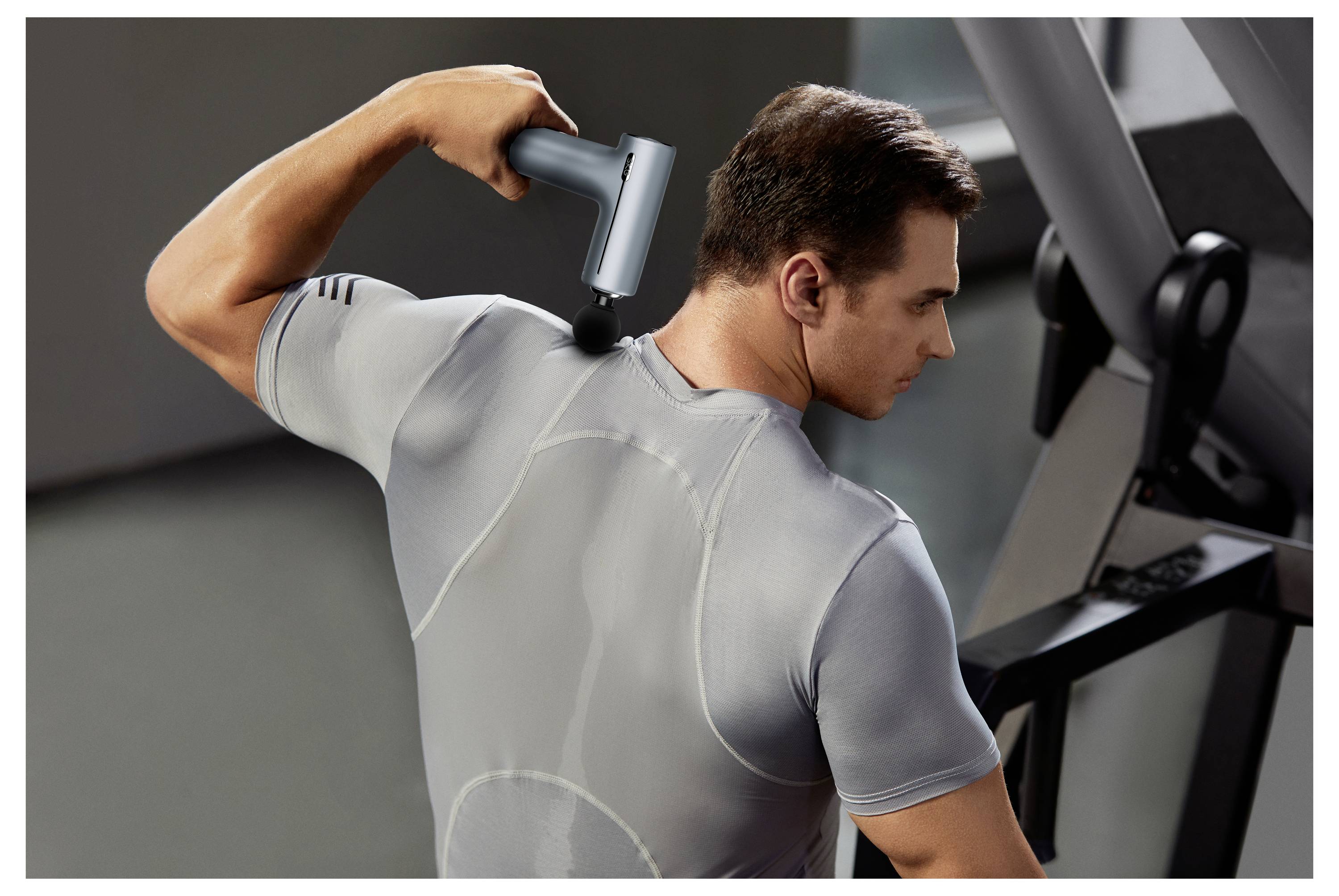 Ein Mann verwendet eine Massagepistole auf seinem oberen Rücken im Fitnessstudio und erholt sich nach einem Workout neben Trainingsgeräten.