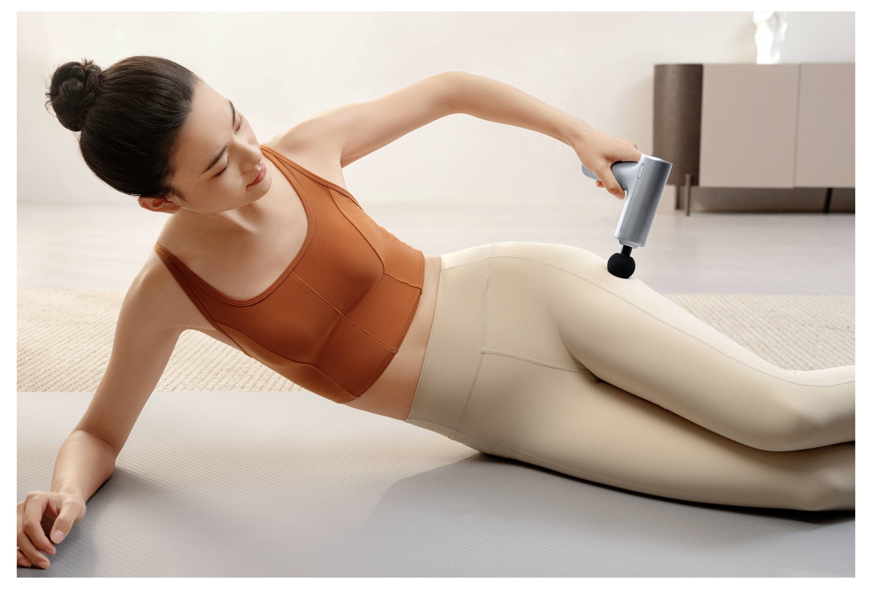 Frau, die ein Massagegerät auf ihrem Oberschenkel verwendet, während sie auf einer Yogamatte in einem hellen Innenraum liegt.