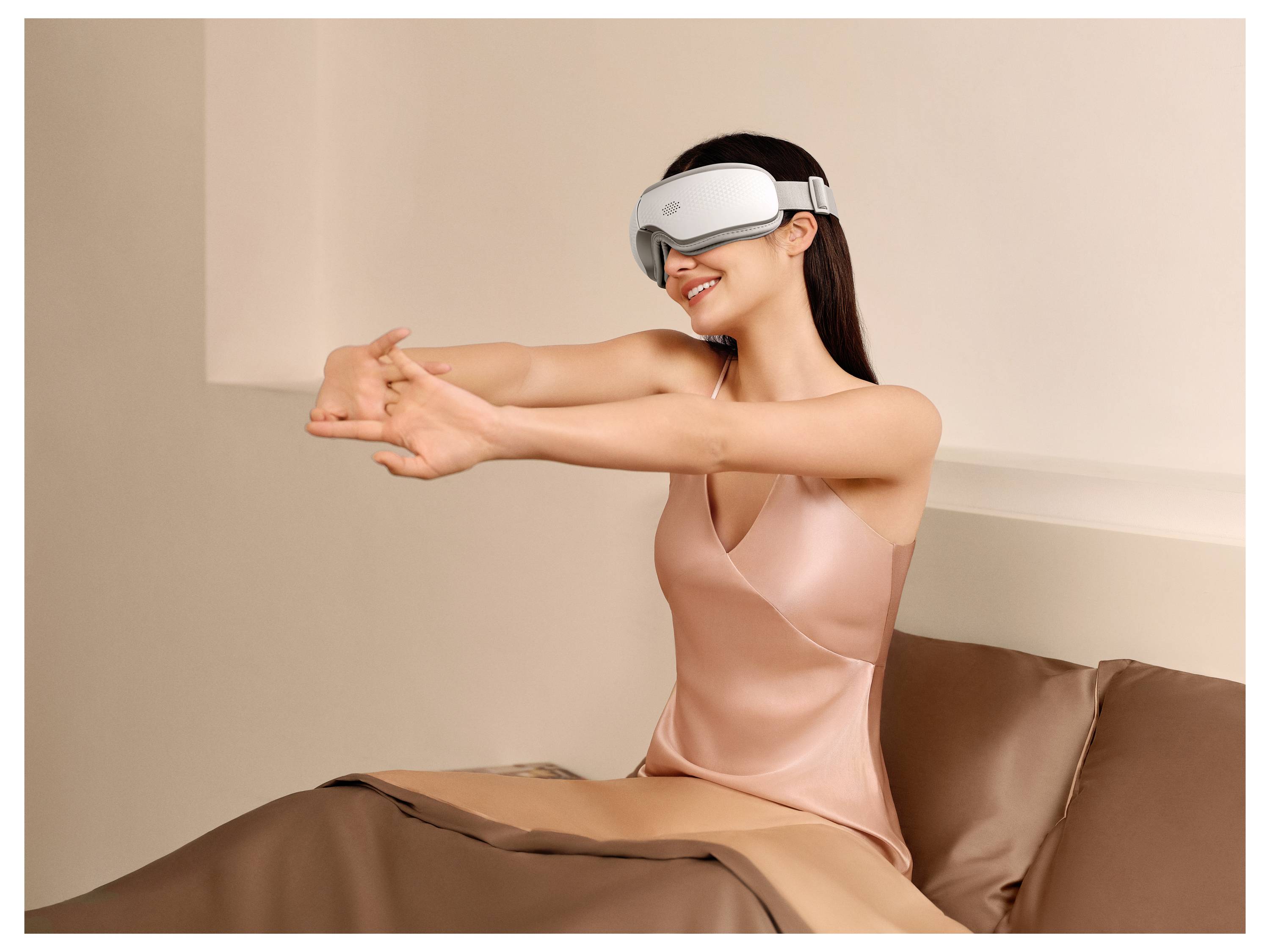 Eine Frau, die eine VR-Brille trägt, sitzt auf einem Bett und streckt ihre Arme nach vorne, während sie in einem sanft beleuchteten Schlafzimmer lächelt.