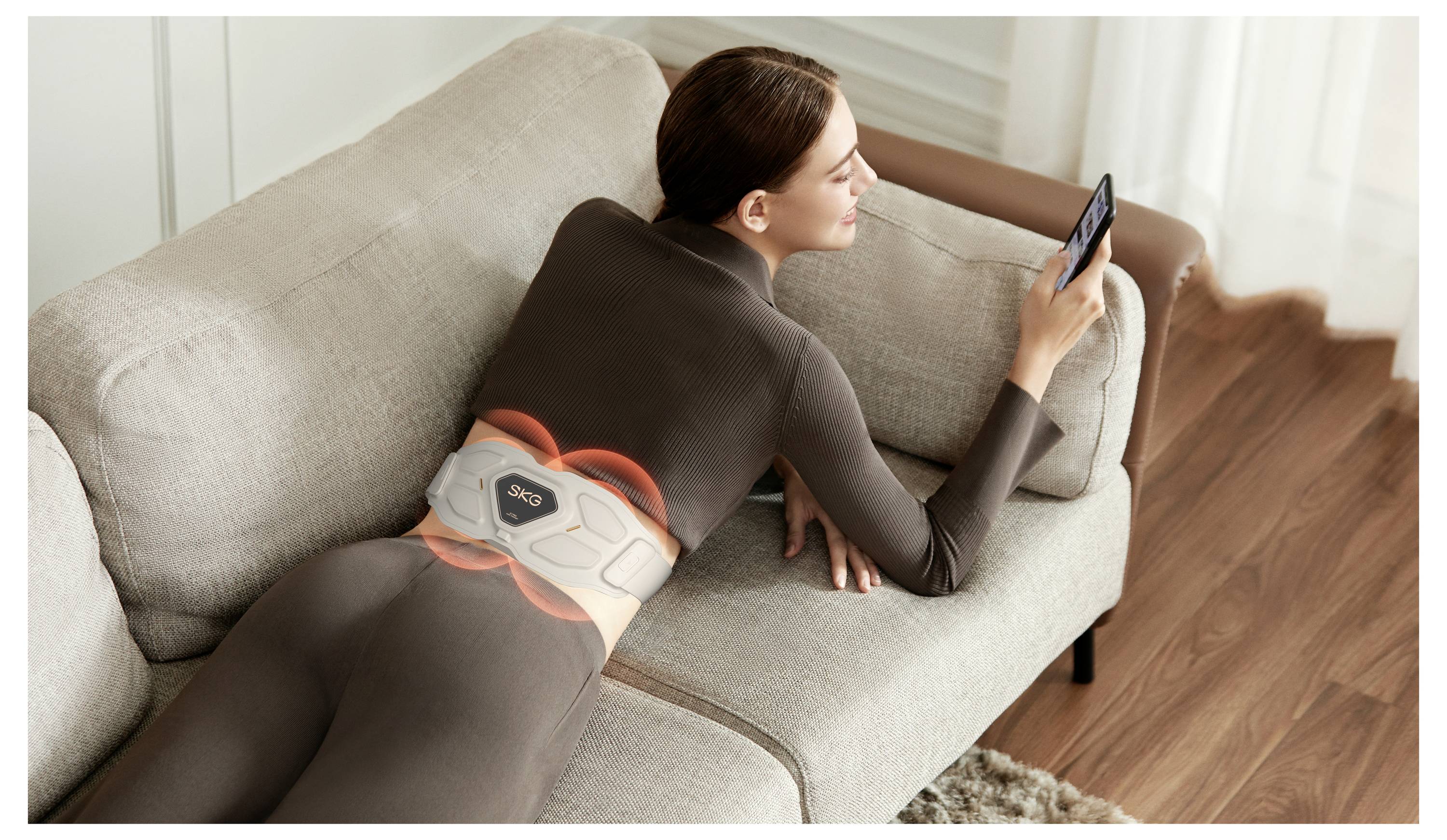 Frau, die auf einem Sofa liegt und ein Smartphone benutzt, während sie einen Lendenwirbel-Massagegürtel trägt, der leuchtet, um Wärme oder Vibration anzuzeigen.