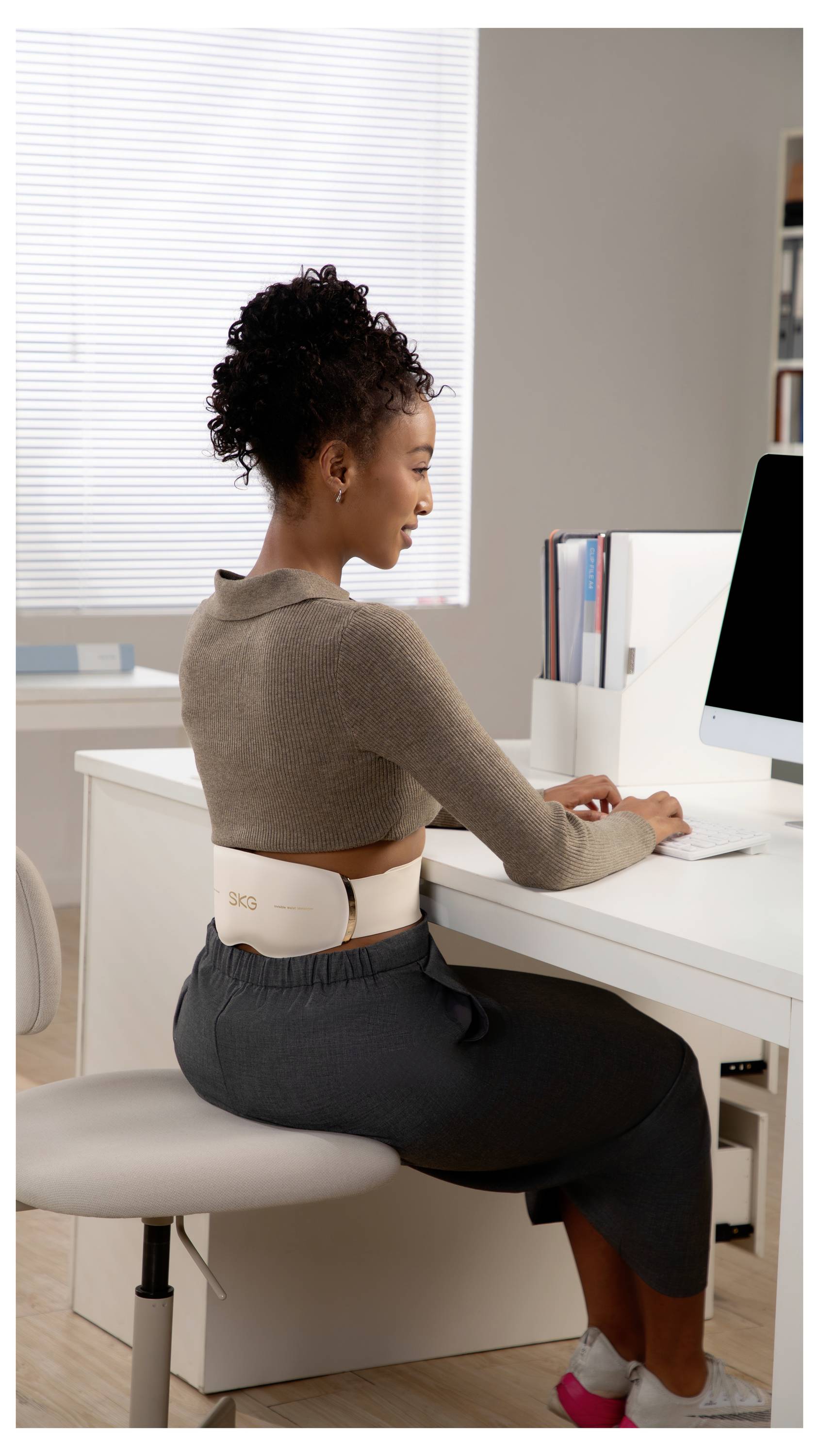 Eine Frau, die einen Rückenstützgürtel trägt, sitzt an einem Schreibtisch und nutzt einen Desktop-Computer in einem Homeoffice.