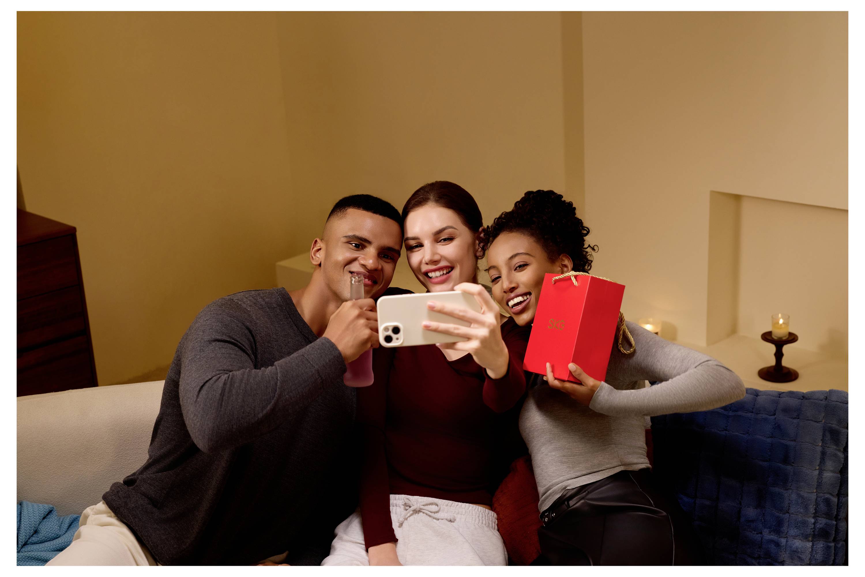Drei Freunde sitzen auf einem Sofa und machen ein Selfie, lächeln und halten ein Telefon und eine rote Geschenkbox in einem gemütlichen Wohnzimmer.