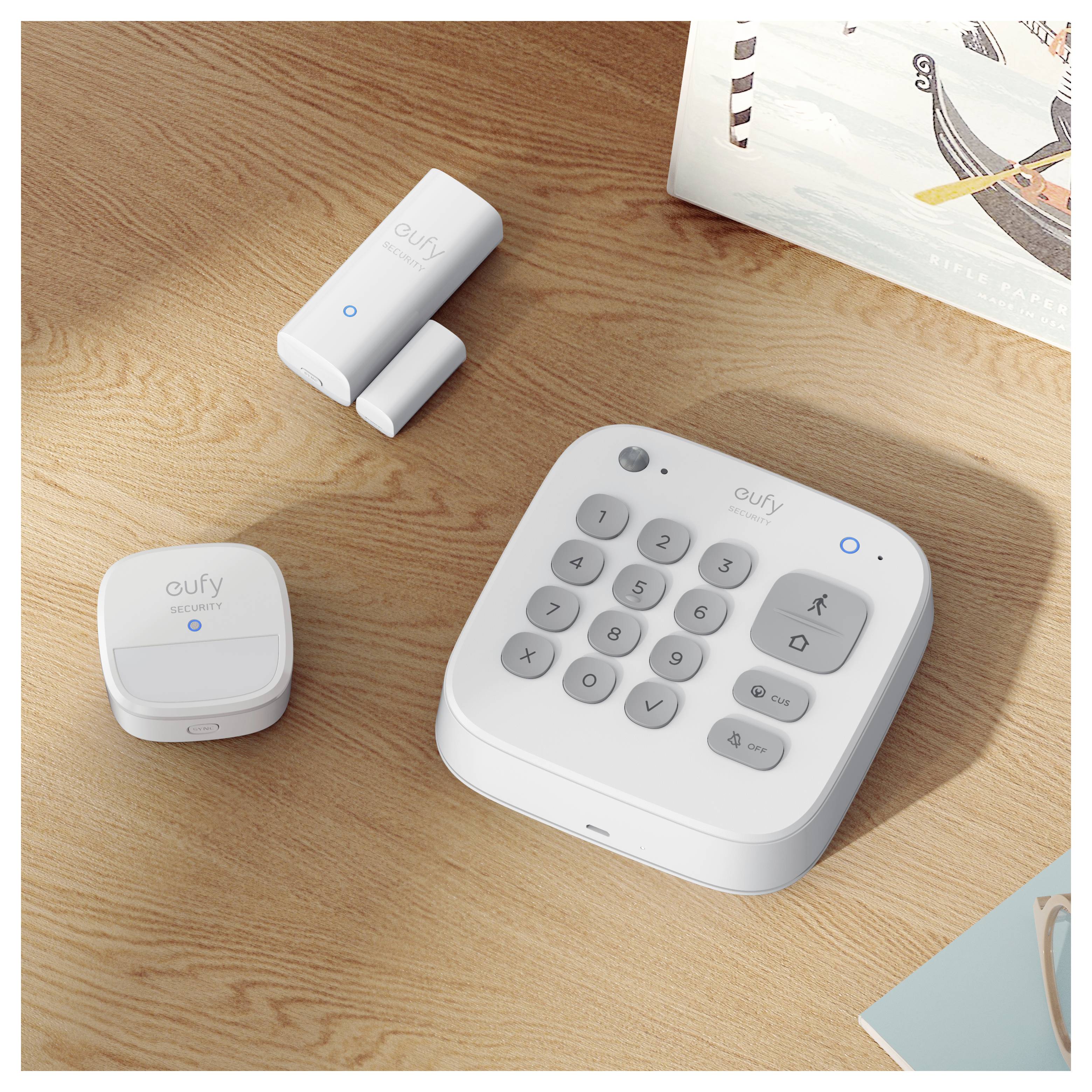 Eufy-Sicherheitsgeräte auf einem Holztisch: ein Keypad-Hub mit Kamera und Tasten sowie ein Sensor und zwei kleine Zubehörteile.