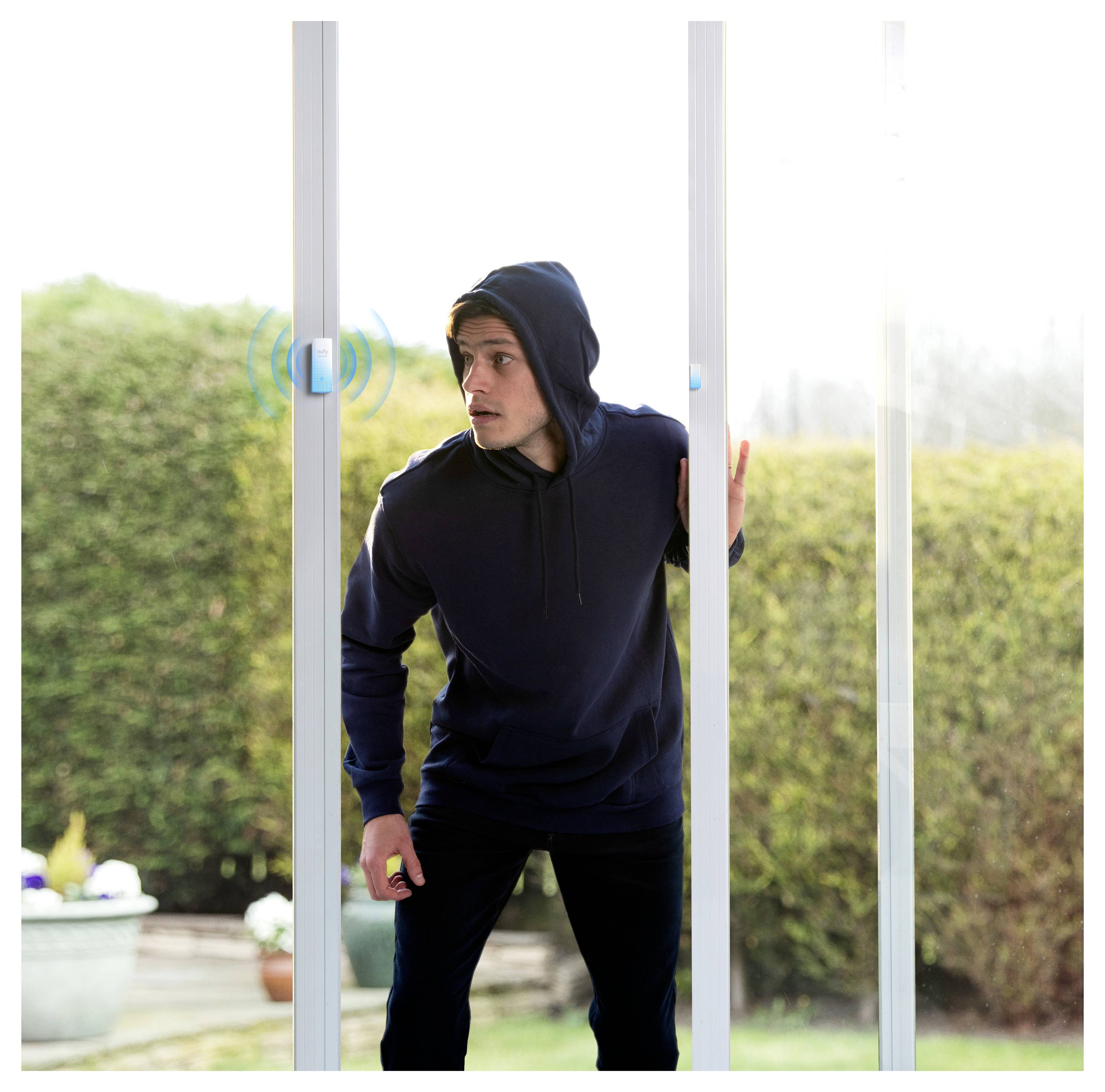 Ein Mann in einem dunklen Kapuzenpullover schaut durch eine Glasschiebetür, als würde er in ein Haus eindringen, mit einem sichtbaren Türgriff in der Nähe.