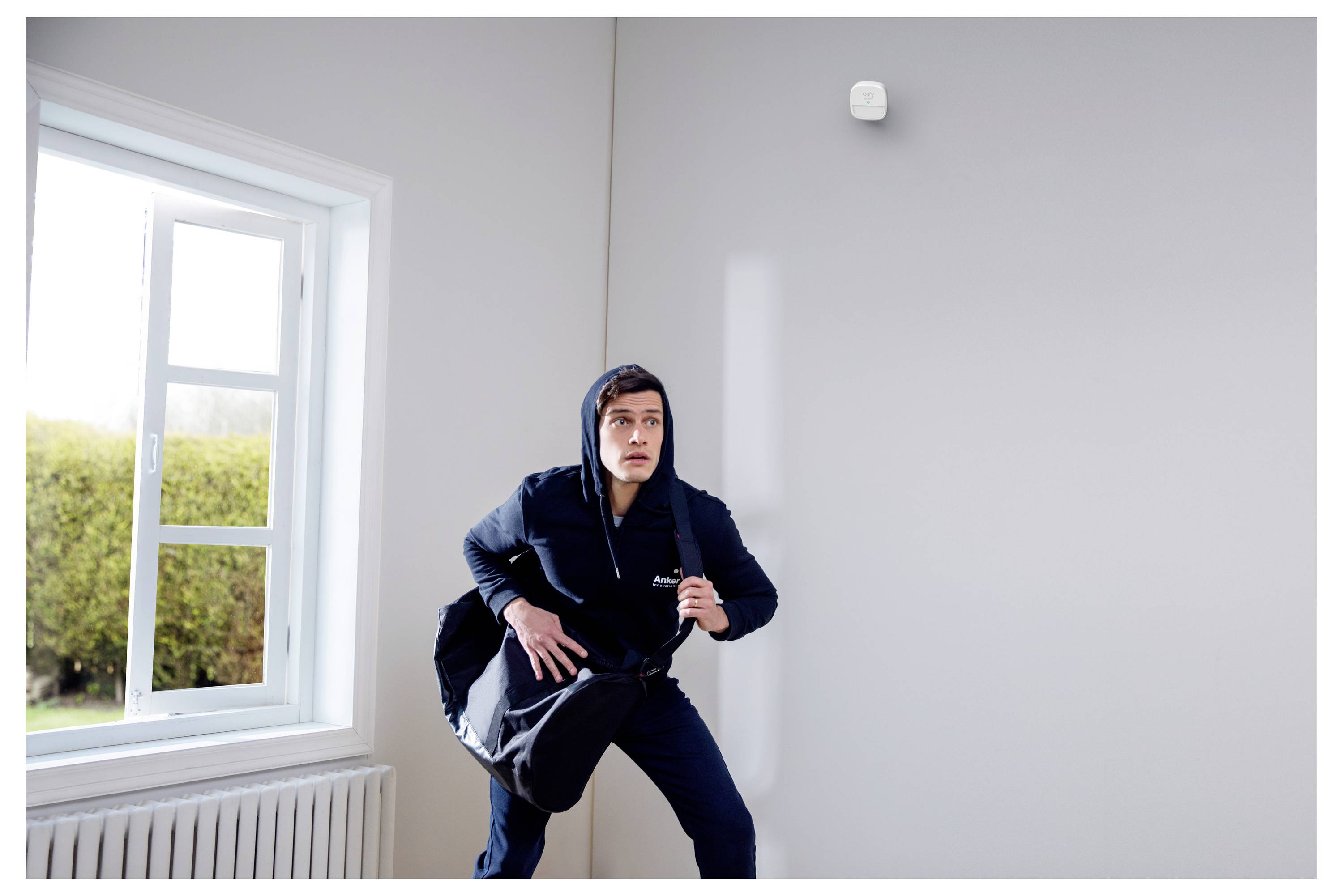 Ein Mann in einer Kapuzenjacke hockt sich an einem Fenster in einem ansonsten leeren Raum hin und hält eine Tasche, als würde er sich verstecken oder etwas bewegen wollen.
