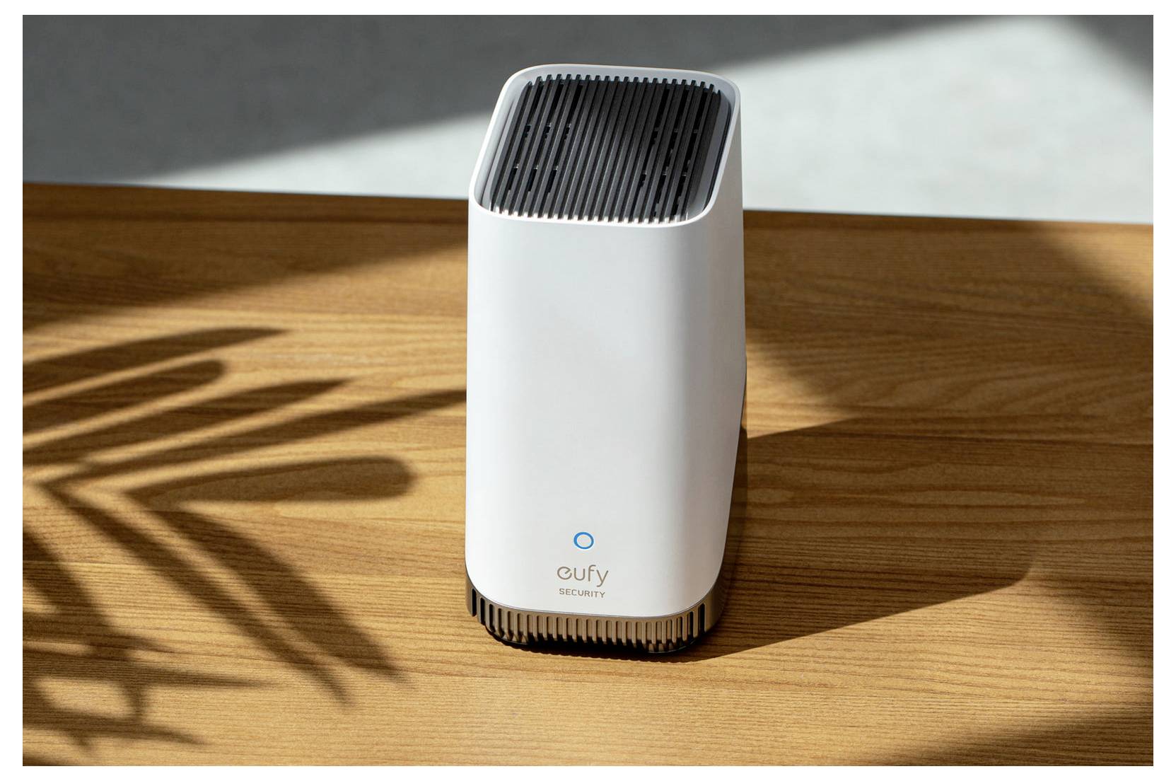 Weißes Eufy Security-Gerät, das auf einem Holztisch in der Sonne steht, mit Pflanzenschatten, die daneben geworfen werden.