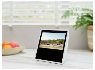 Ein digitaler Bilderrahmen auf einem weißen Tisch zeigt eine Landschaftsszene an, mit Obst, einer Pflanze und Jalousien im Hintergrund.