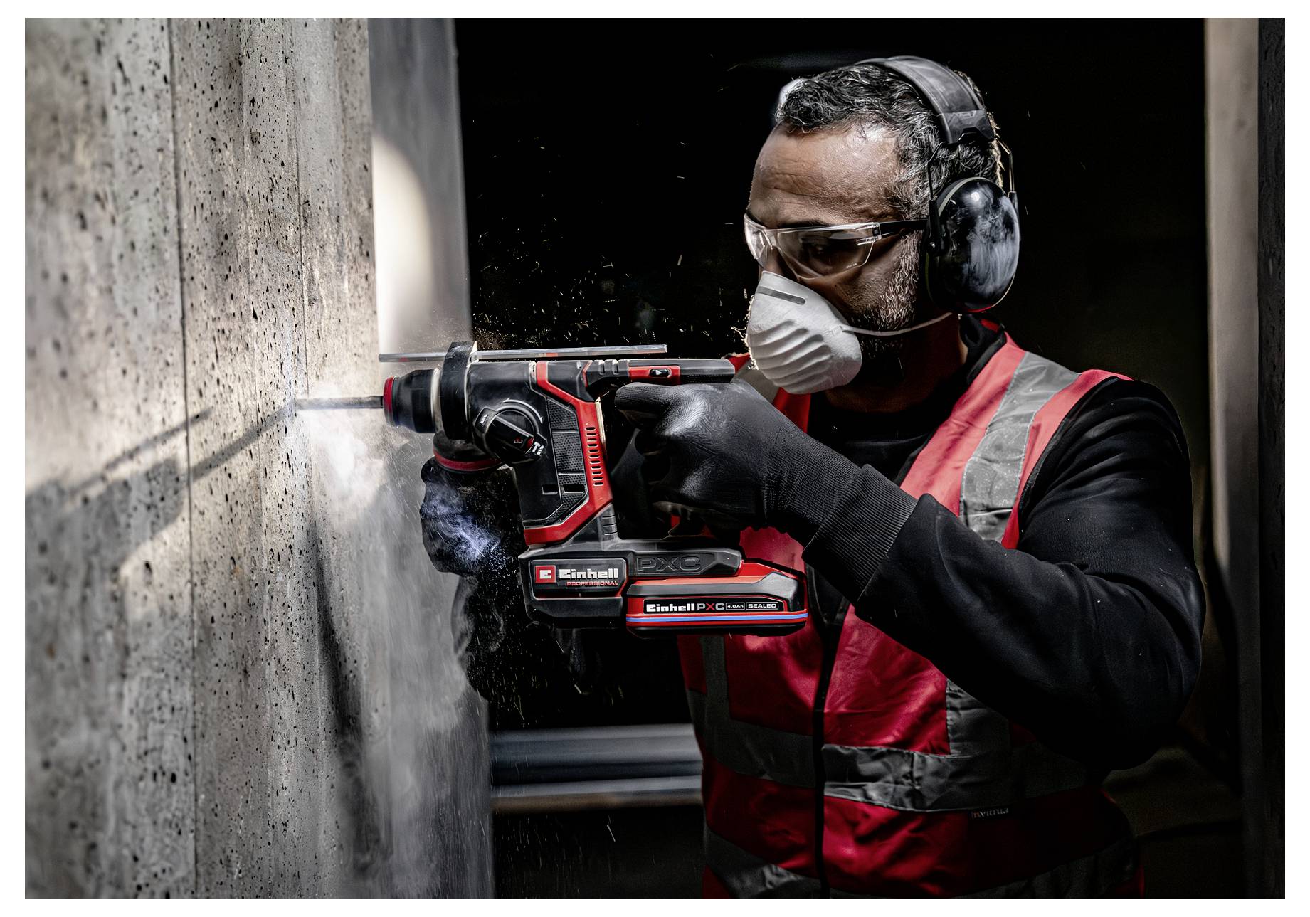 Bauarbeiter, der mit einem kabellosen Bohrhammer in eine Betonwand bohrt, trägt Schutzbrille, Maske, Handschuhe und Gehörschutz.