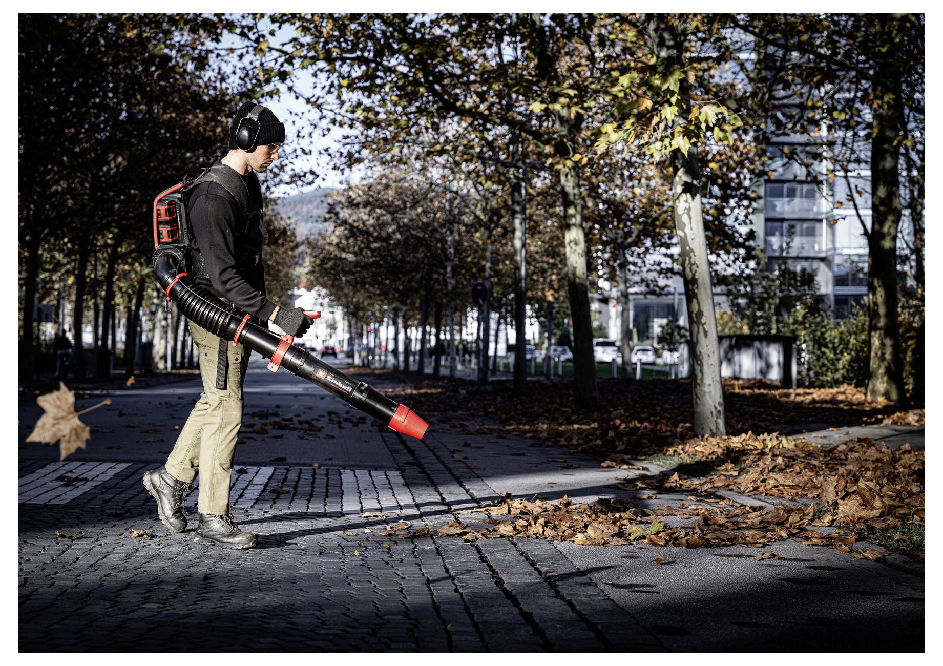 Ein Arbeiter, der ein Rucksack-Laubgebläse verwendet, um herabgefallene Blätter von einem baumgesäumten Gehweg im Herbst zu entfernen.