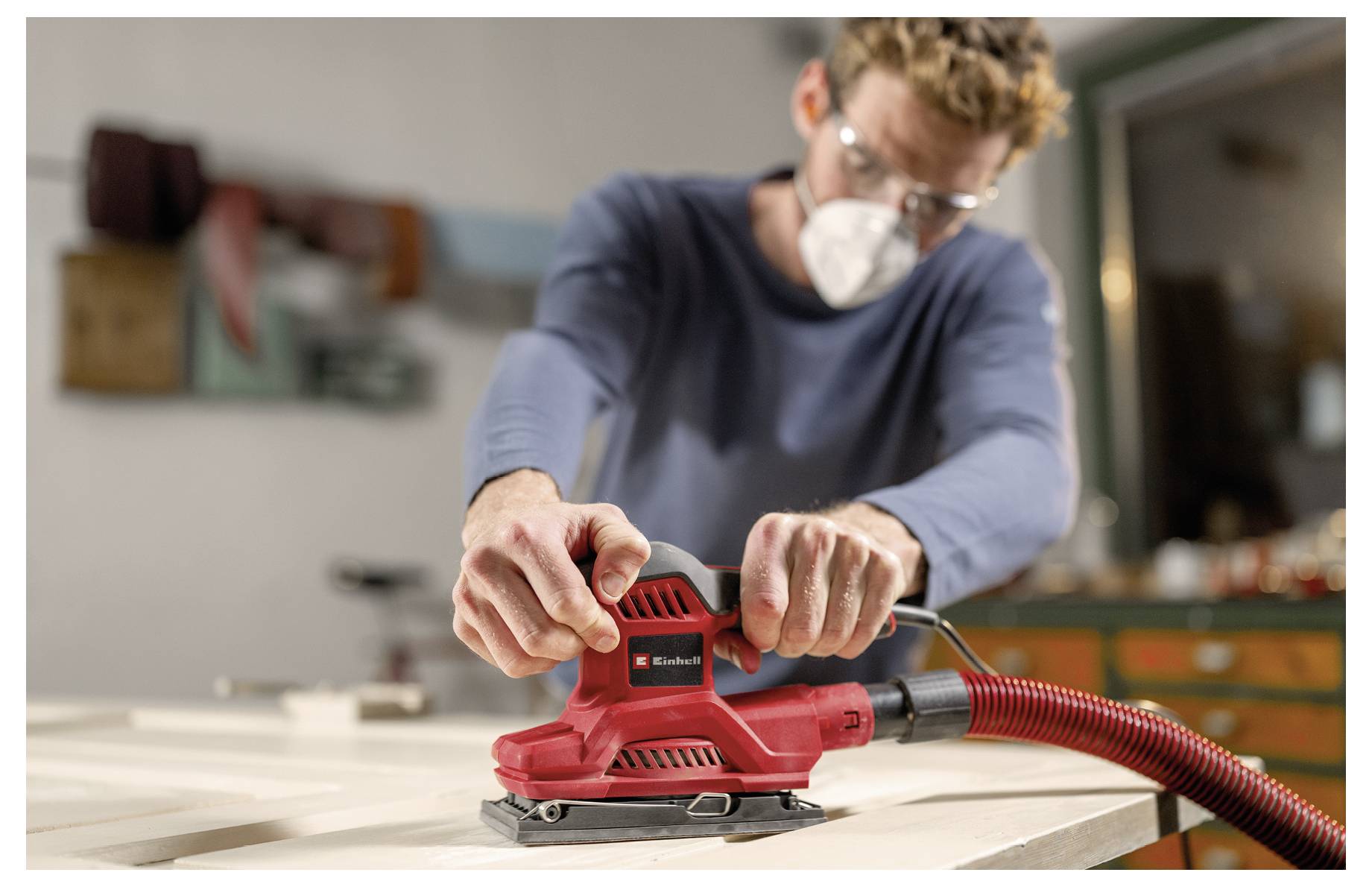 Ein maskierter Mann schleifen ein Holzbrett mit einem roten elektrischen Elektroschleifen in einer Werkstatt.