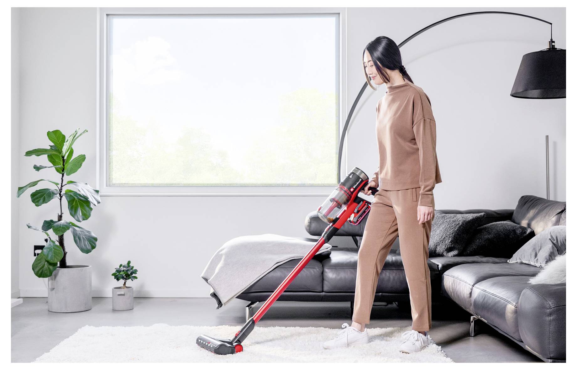 Frau staubsaugt einen weißen Teppich in einem hellen Wohnzimmer neben einem schwarzen Sofa und einer Stehlampe.